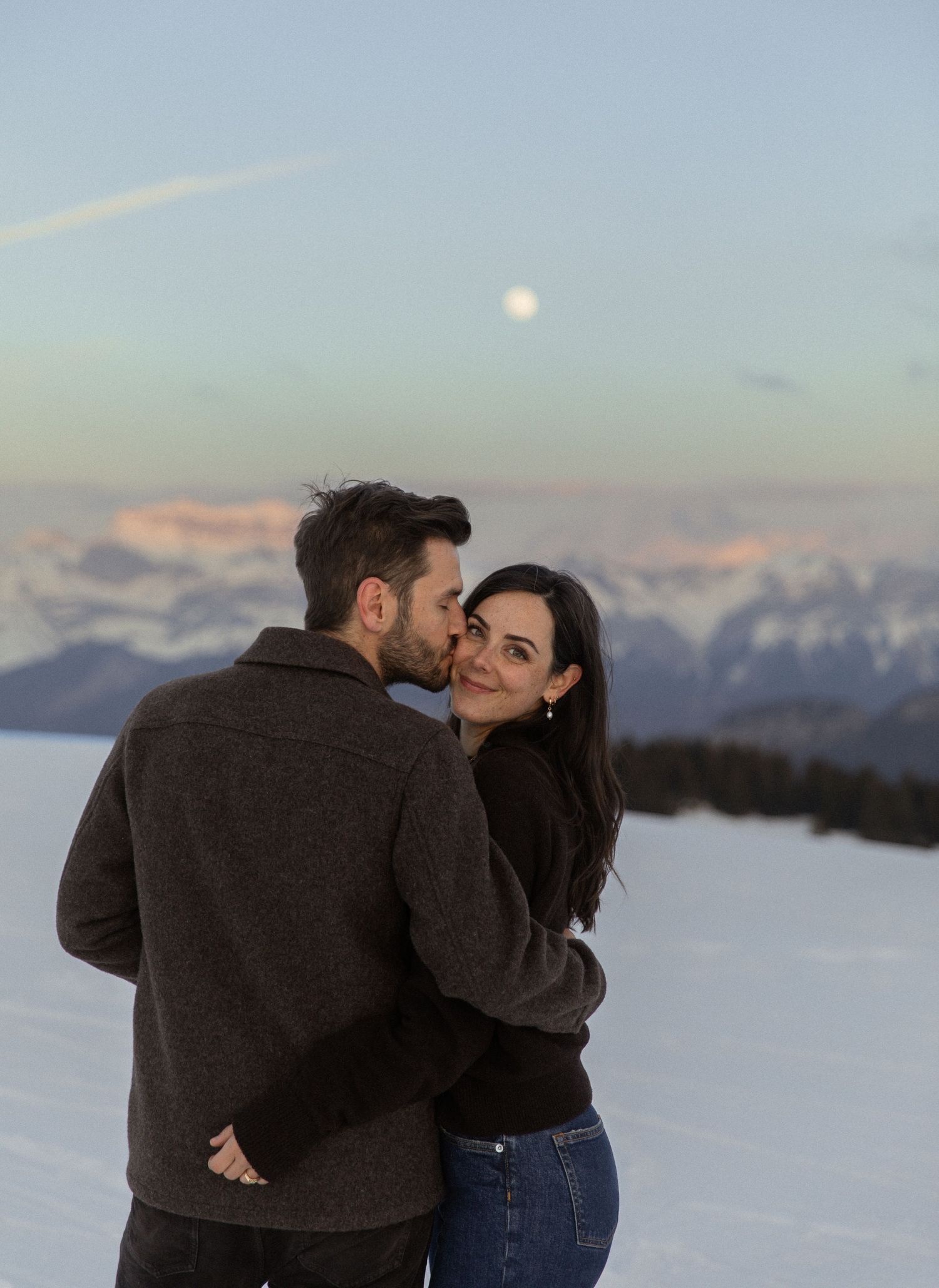 A couple shares an intimate moment in the snowy mountains at sunset with snow-capped peaks in the background.