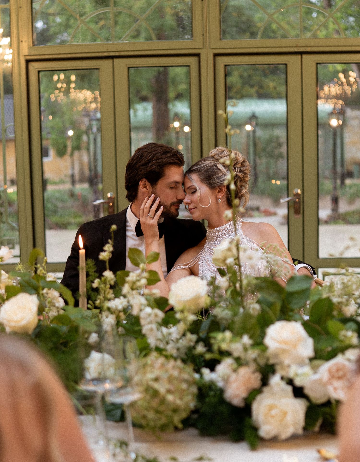Bride and groom share a romantic moment at elegantly decorated wedding reception table with white floral arrangements.