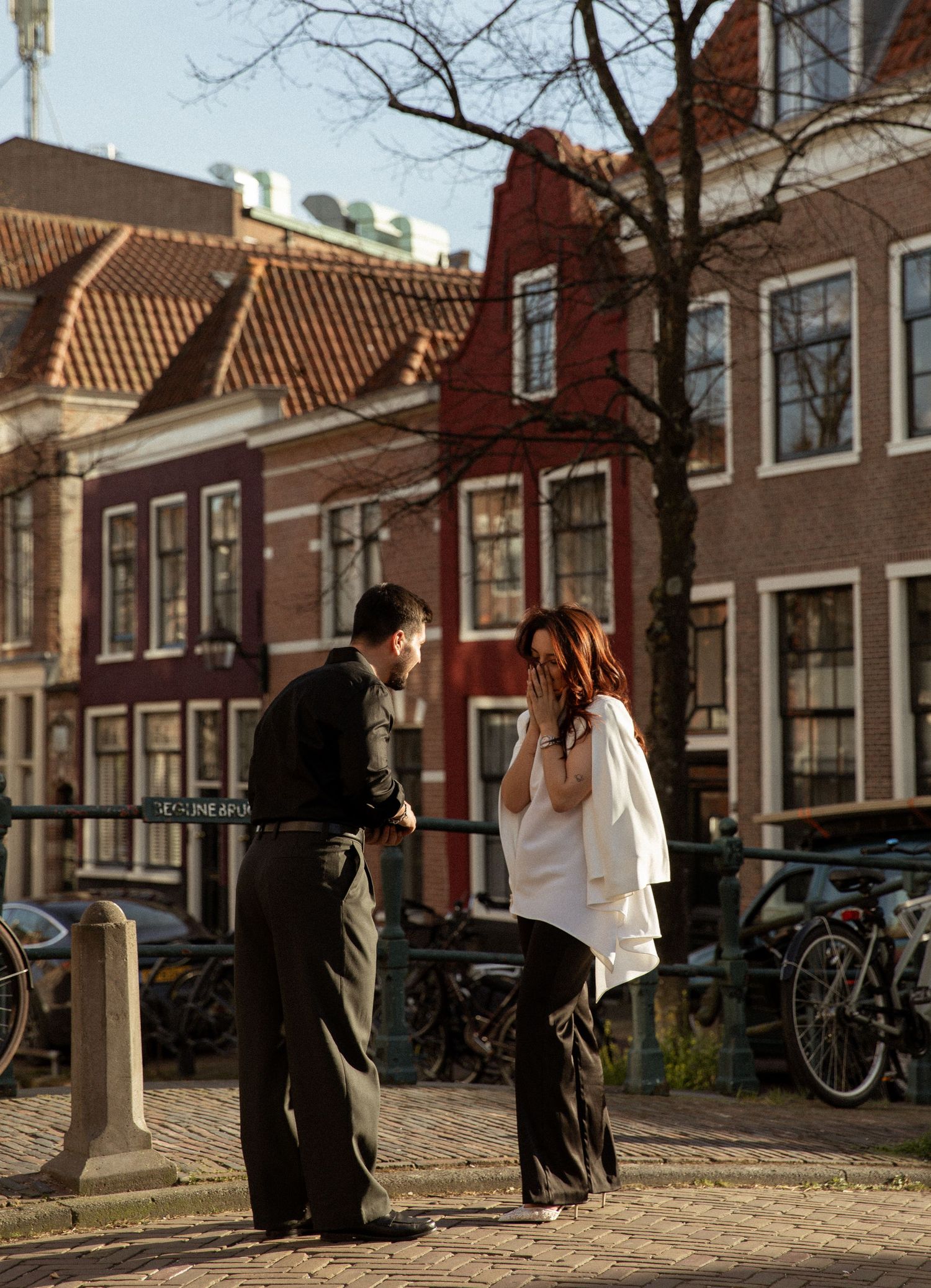 Couple sharing a romantic moment on a sunny Amsterdam canal street, surrounded by classic Dutch architecture and bicycles.