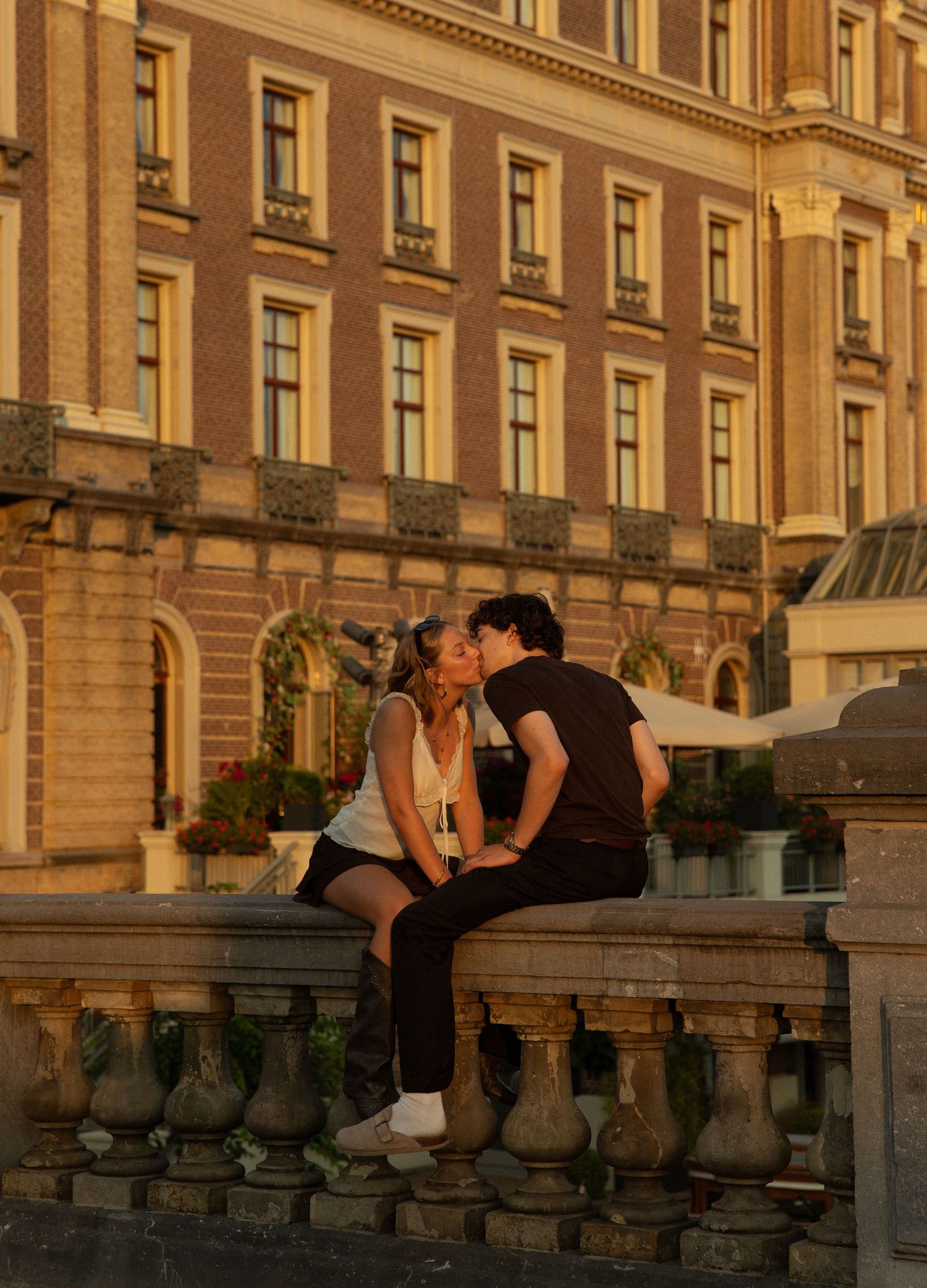 Couple sharing a romantic kiss at sunset on a stone balustrade in front of a grand European building.