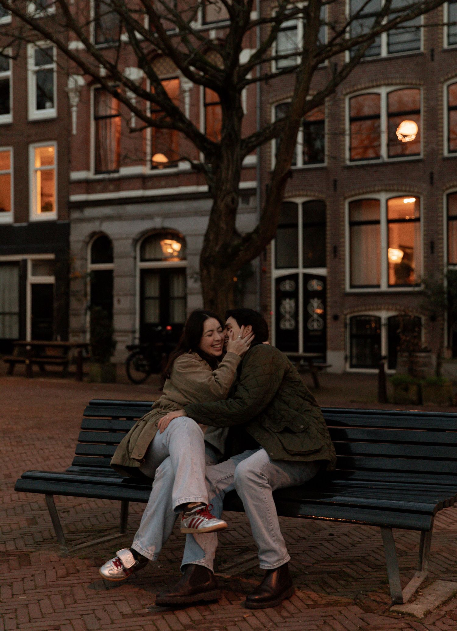 Couple sharing tender moments on a park bench at dusk with city buildings in the background.