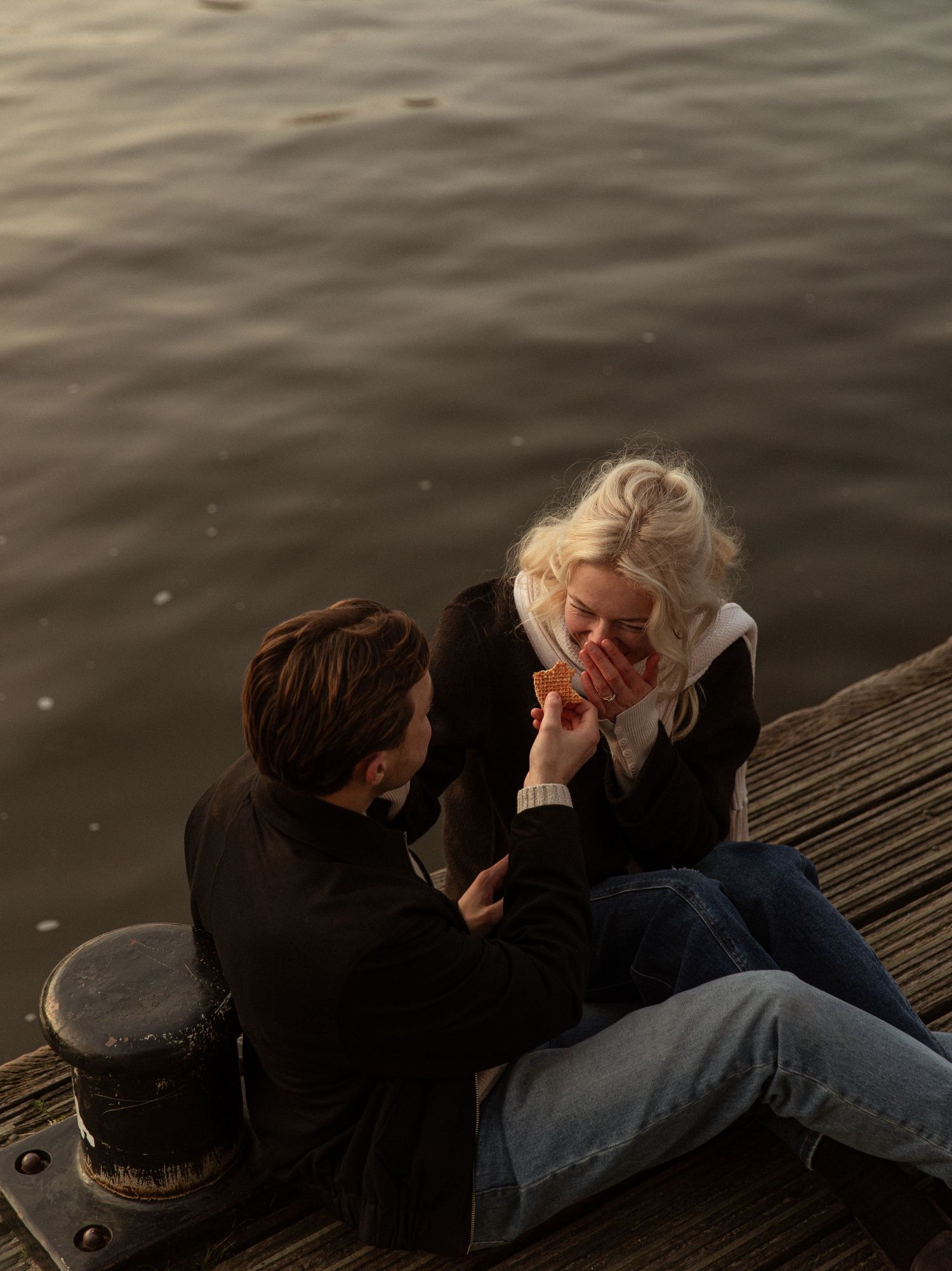 Two people sit close together on a wooden dock by dark rippling water at dusk.