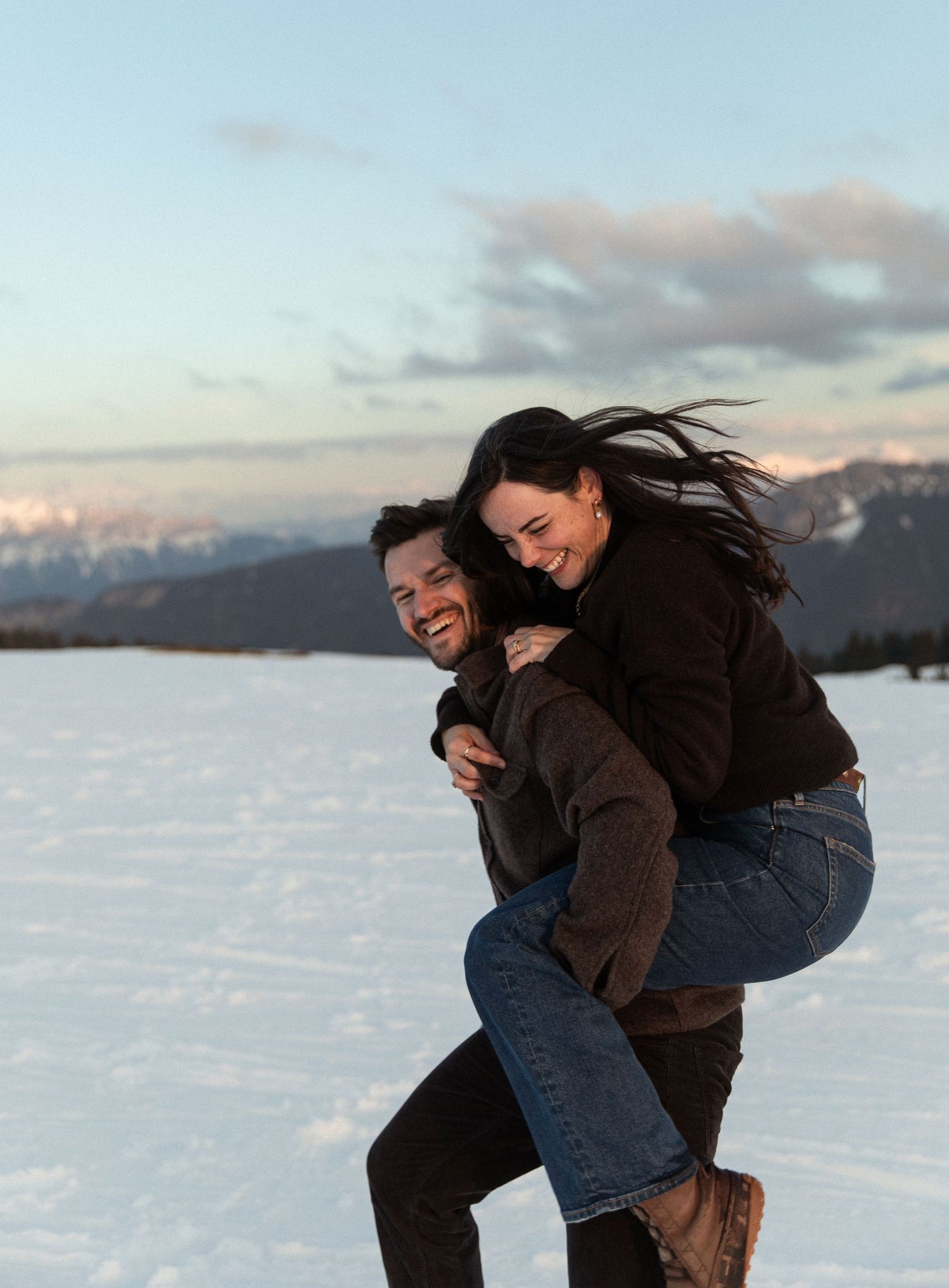 Two people share a playful moment together in the snow with snow-capped mountains visible in the background at sunset.