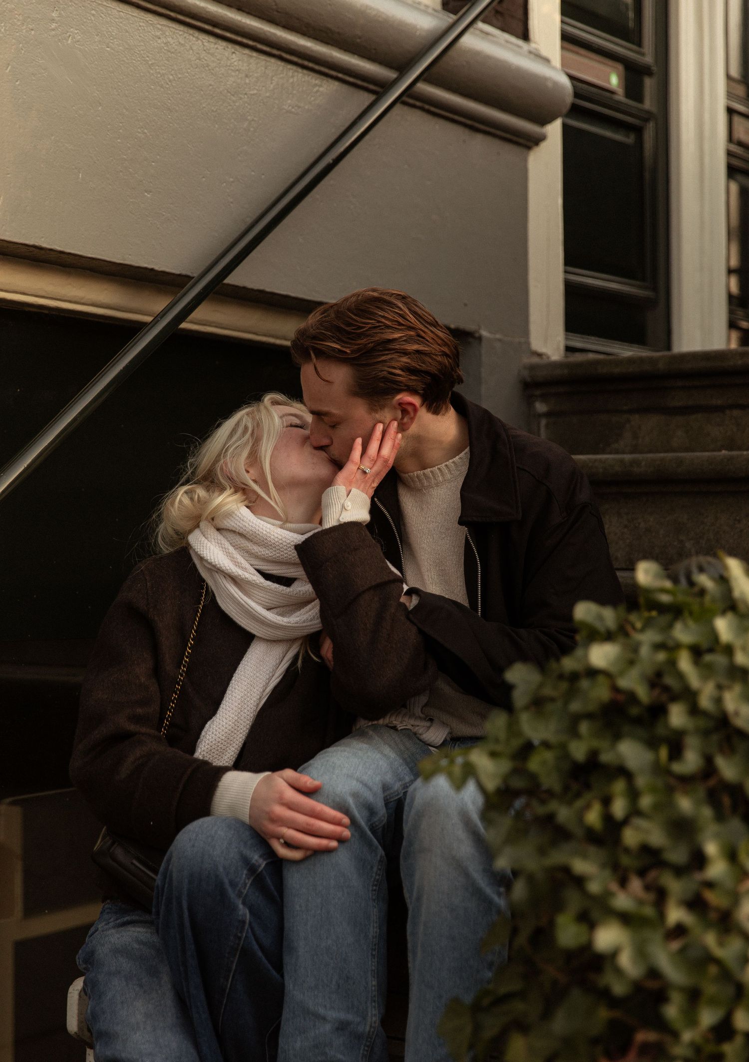A person in jeans and a brown jacket sits alone on steps, appearing distressed with their head in their hands.