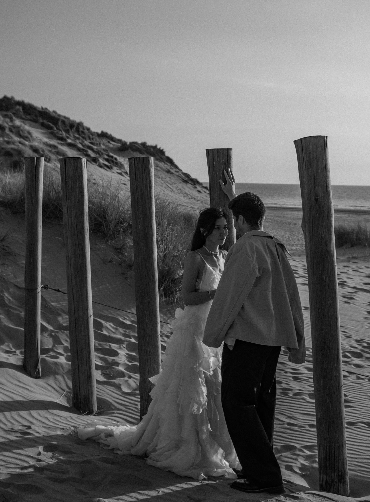 Black and white photo of a bride and groom sharing an intimate moment on a sandy beach surrounded by wooden posts.