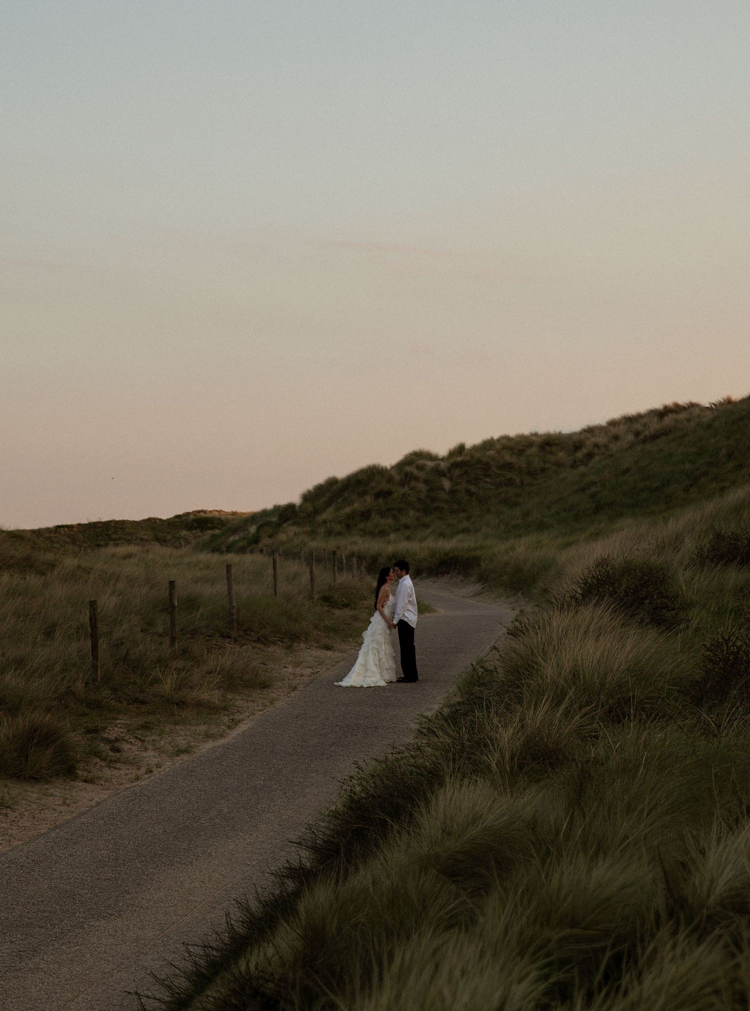 A bride and groom stand together on a winding coastal path surrounded by grass dunes at sunset.