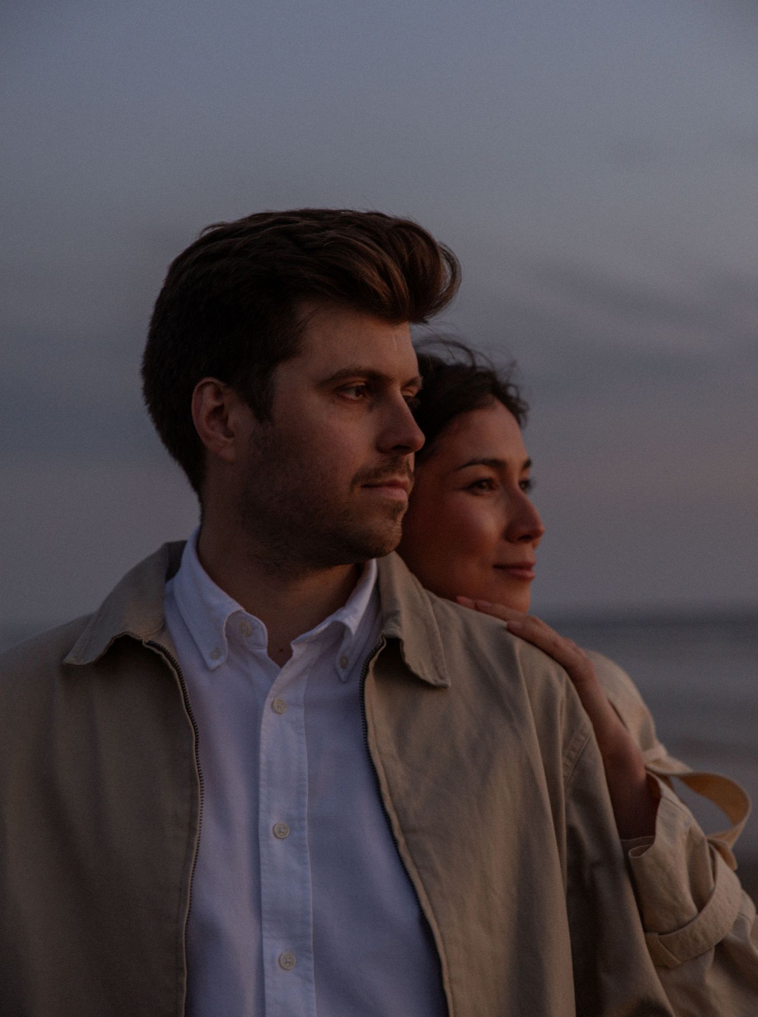 A couple stands together at sunset by the ocean, the man in a beige jacket gazing thoughtfully into the distance.