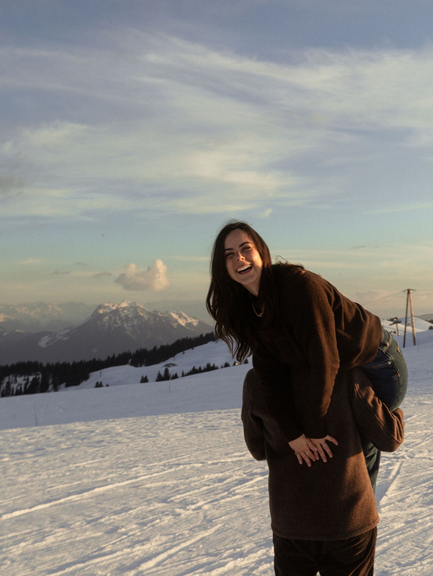A person in a brown jacket laughs while standing in a snowy mountain landscape during sunset.