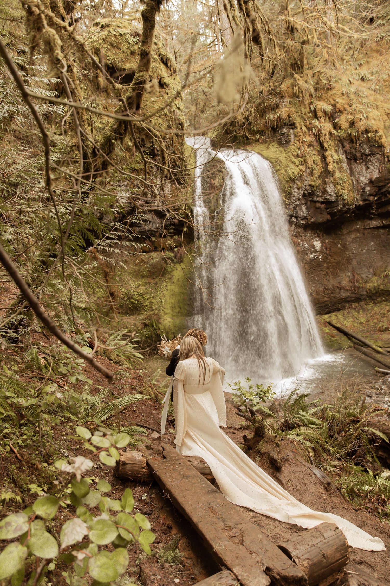 Stunning waterfall creates dramatic backdrop for wedding dress trailing along forest path.