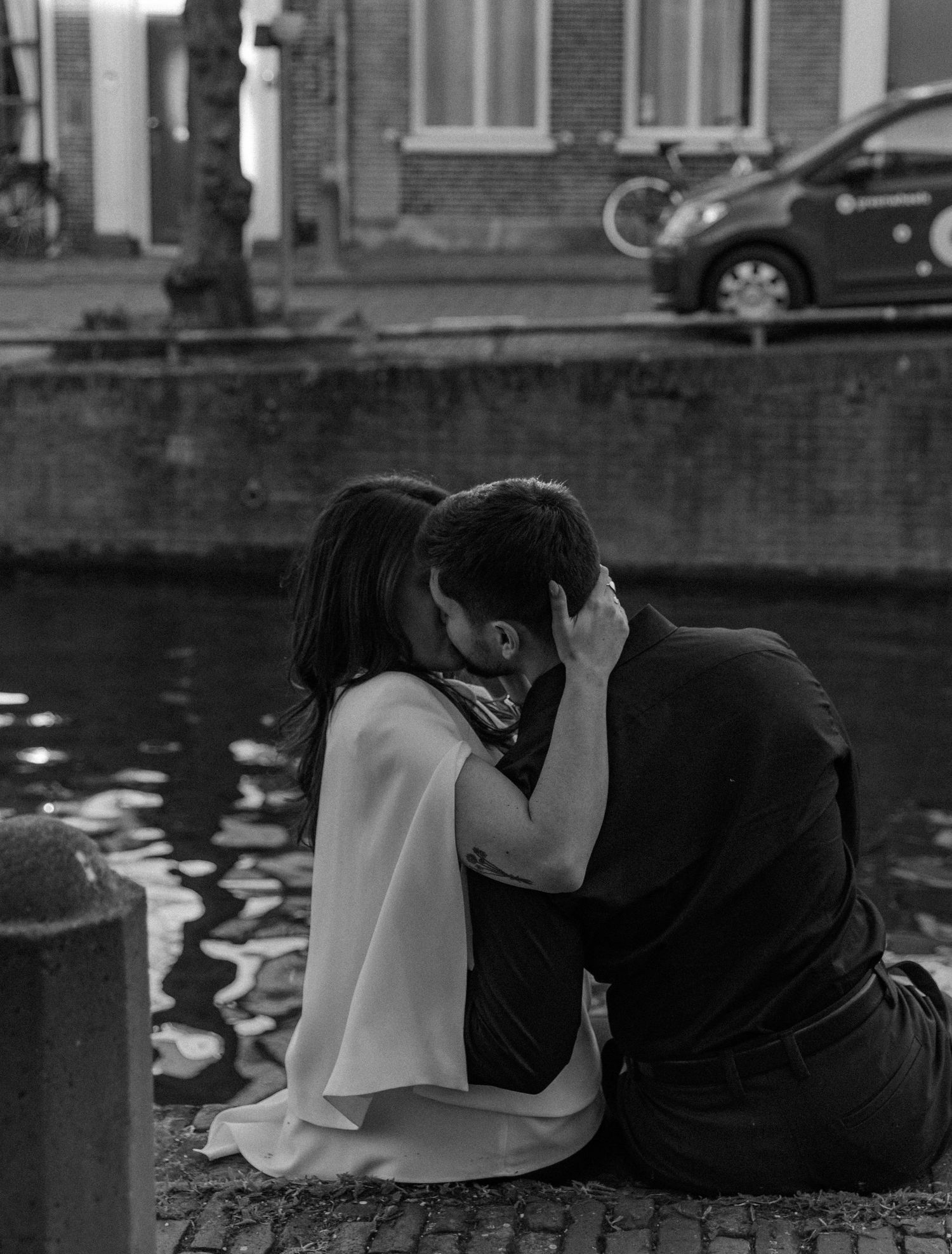 Black and white photo of a couple sharing a romantic kiss beside a canal in a European city.
