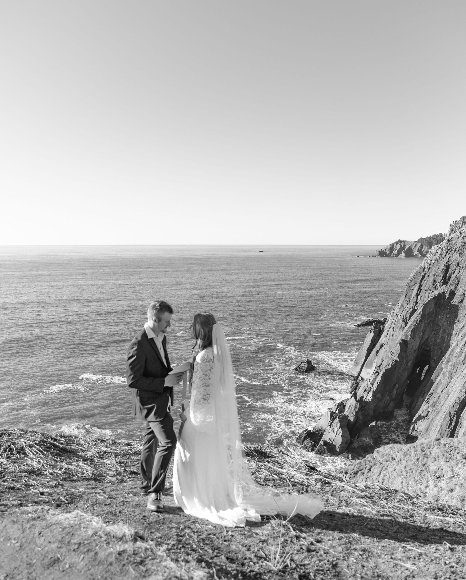 A couple stands together on dramatic coastal cliffs overlooking the ocean waves crashing against rocky shores.