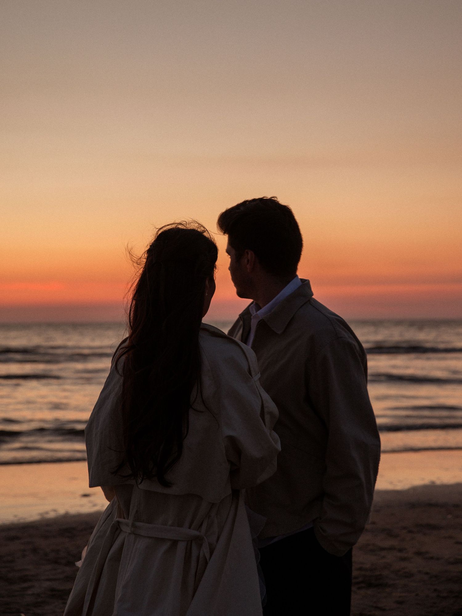 Couple silhouetted against a vibrant ocean sunset, standing close together on a beach at dusk.