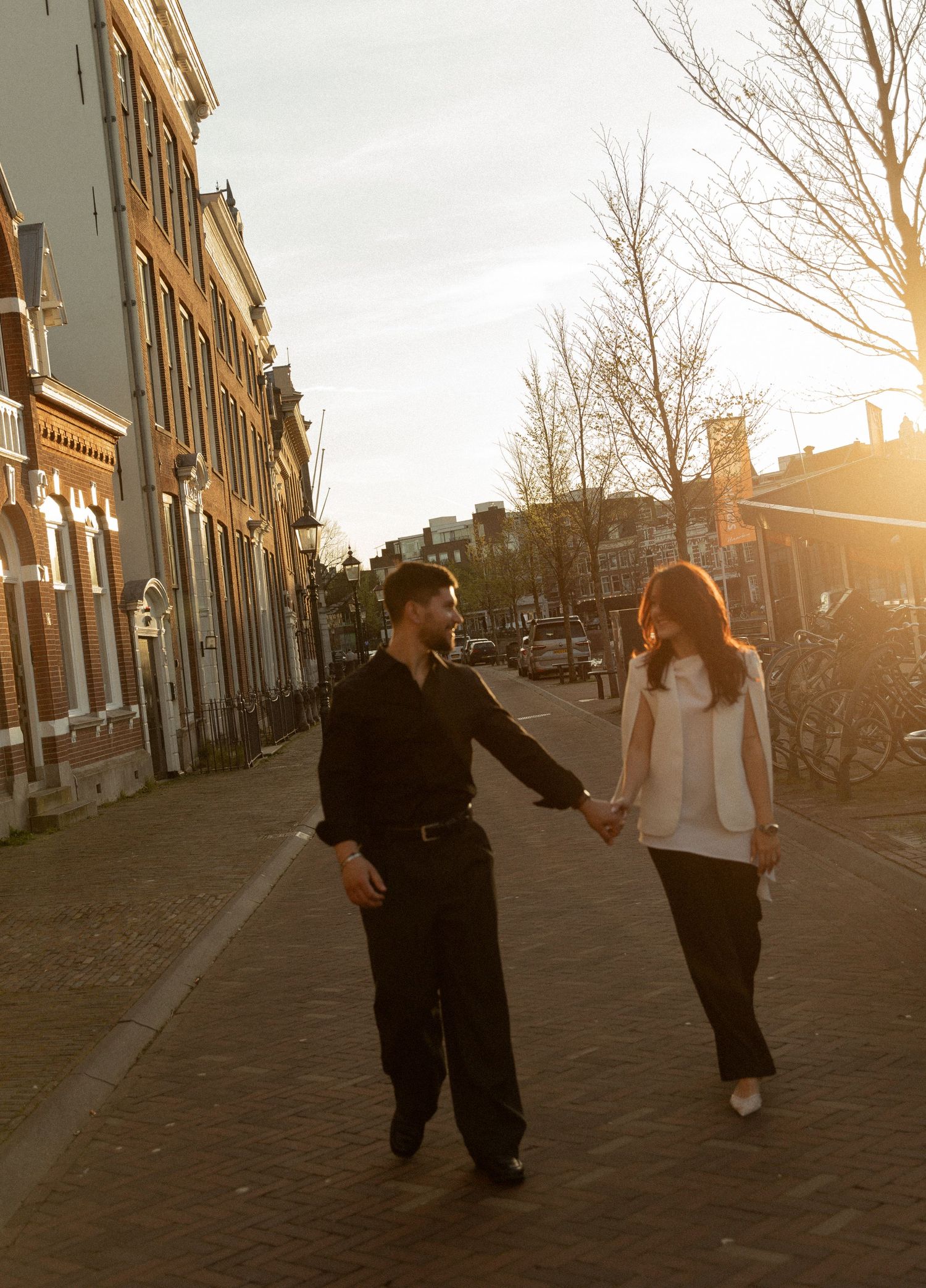 Couple holding hands walking on a sunlit cobblestone street in Amsterdam during golden hour sunset.