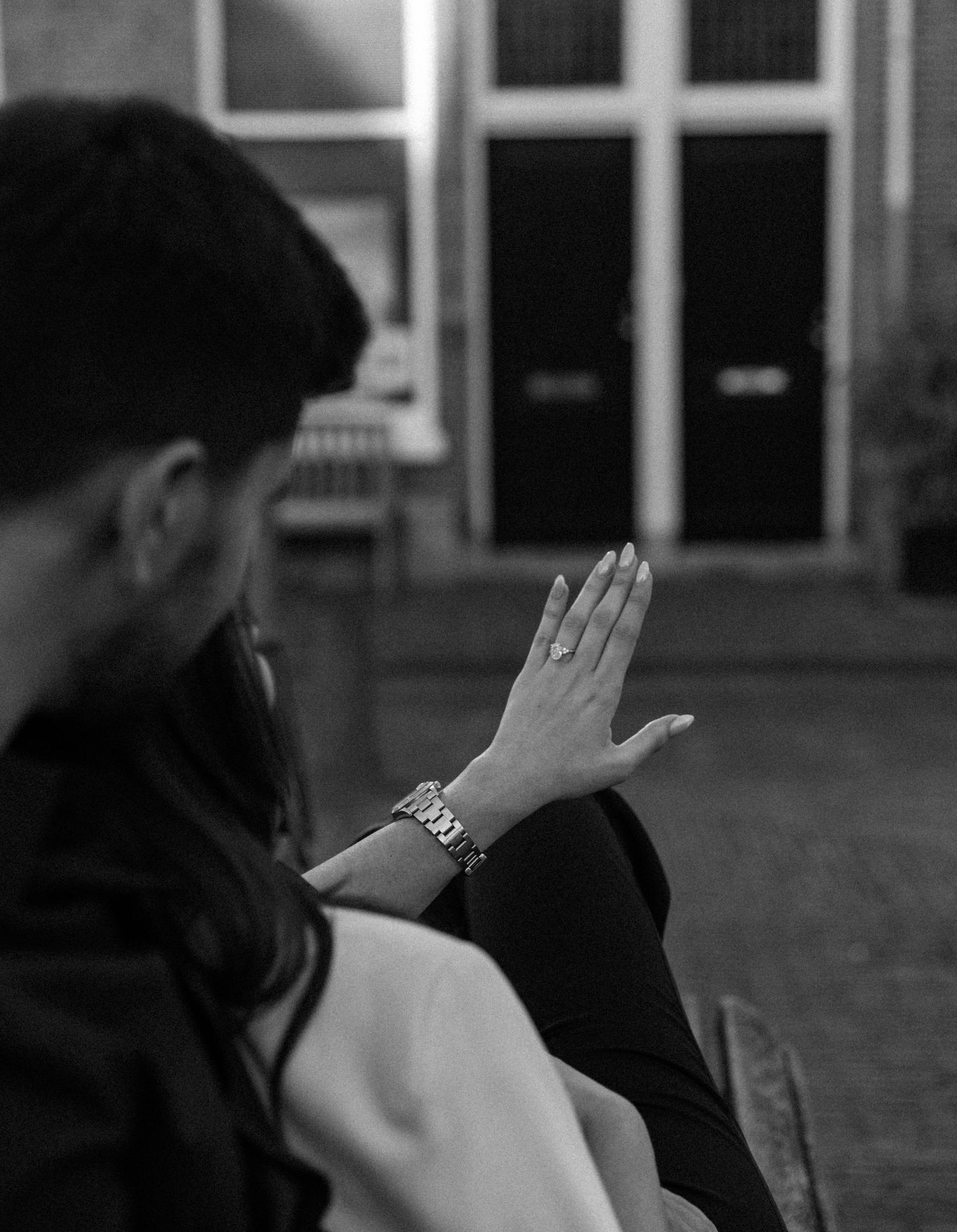Black and white photo of a person gesturing with their hand, wearing a bracelet, seated outdoors near a building.
