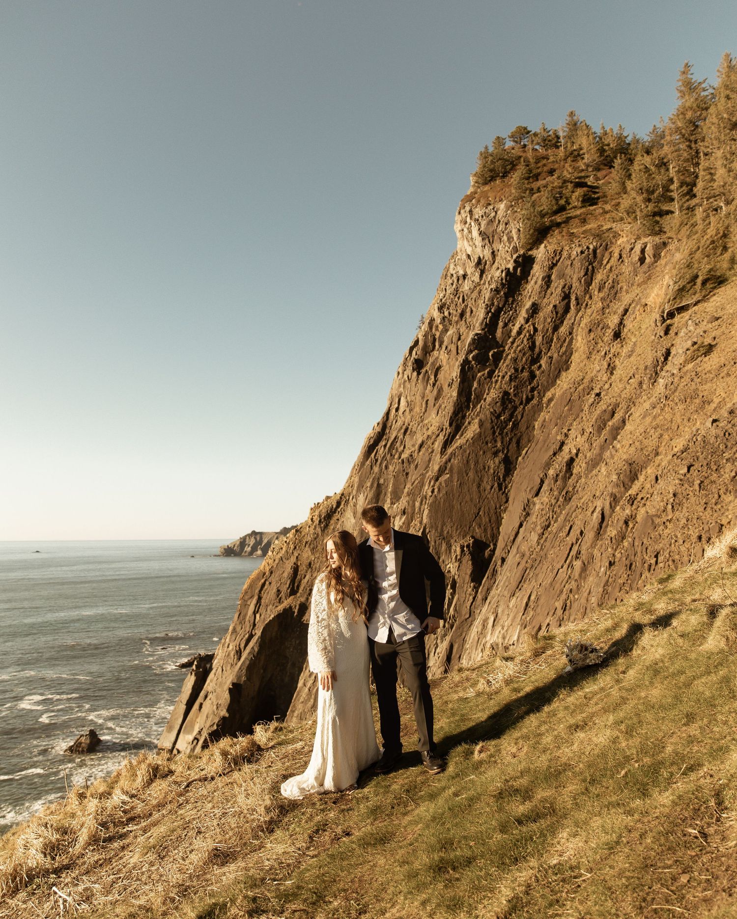 A couple stands on a coastal cliff edge with dramatic rocky cliffs and ocean views in the background.