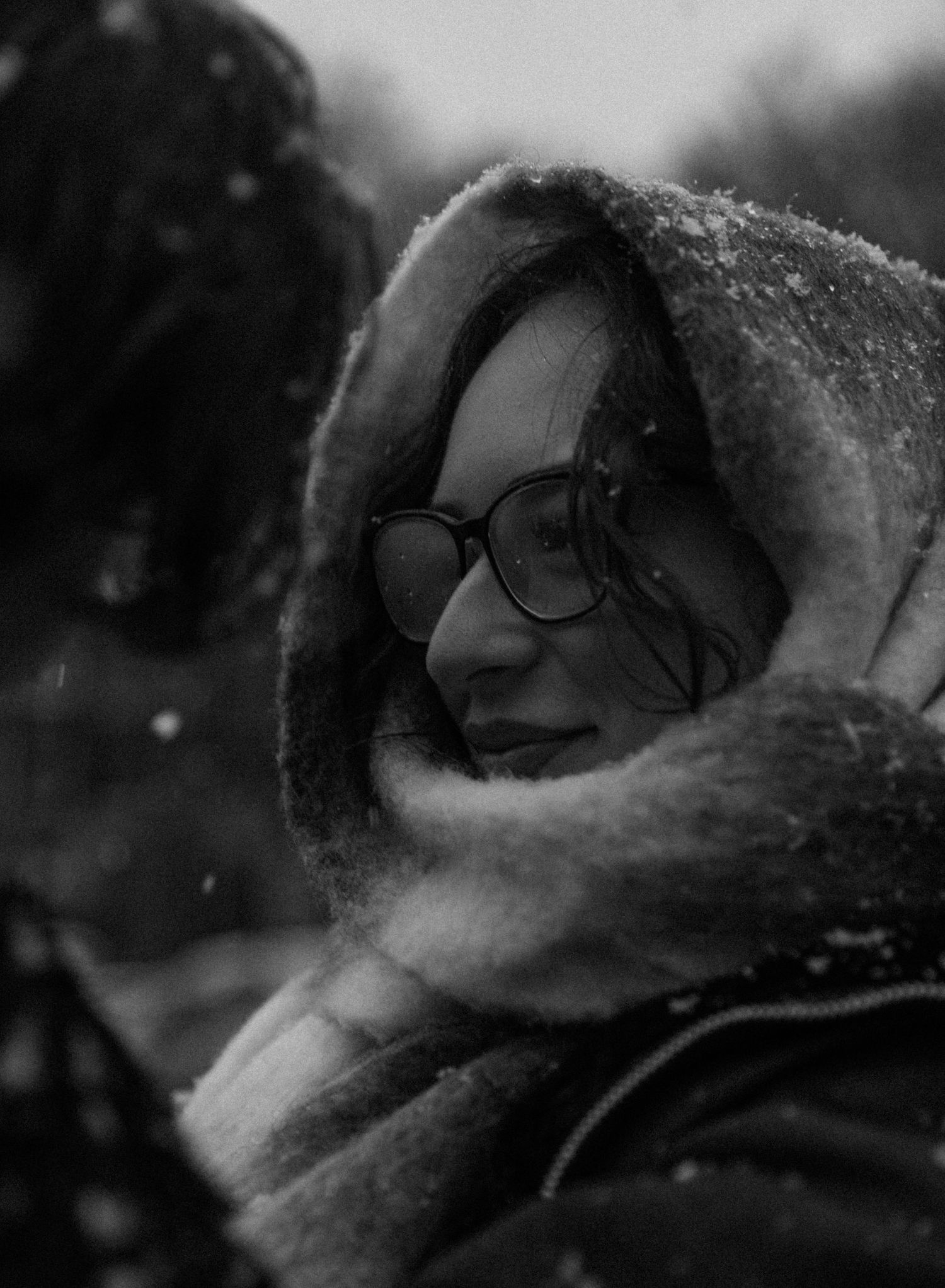 Close-up black and white photo of someone smiling through falling snow.