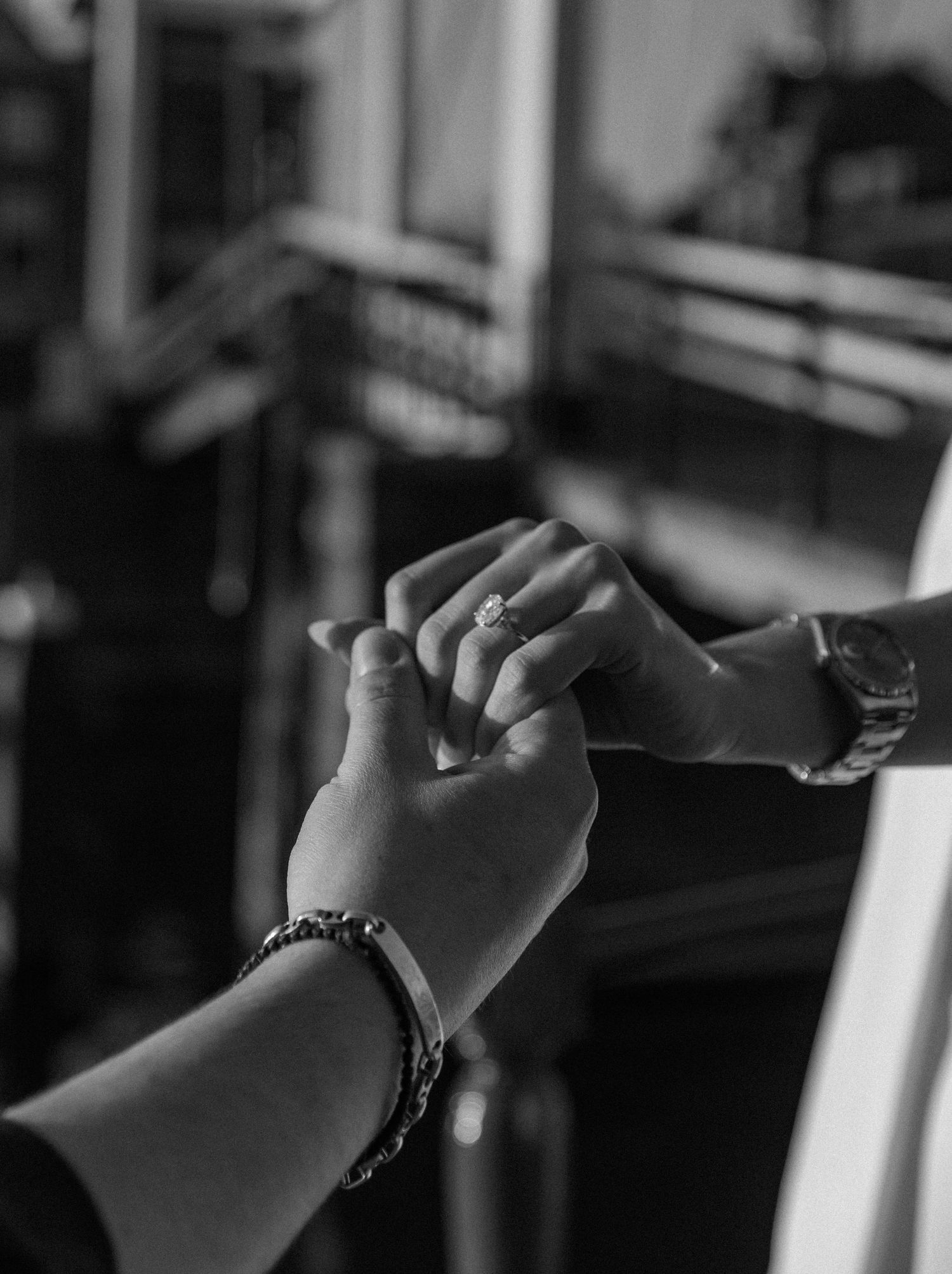 Black and white photo of two hands holding, one wearing a diamond ring and bracelet, the other a watch.