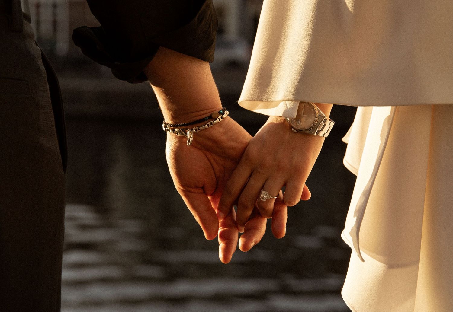 Close-up of couple holding hands, bride wearing diamond ring and bracelet, groom in dark suit, golden sunset light.