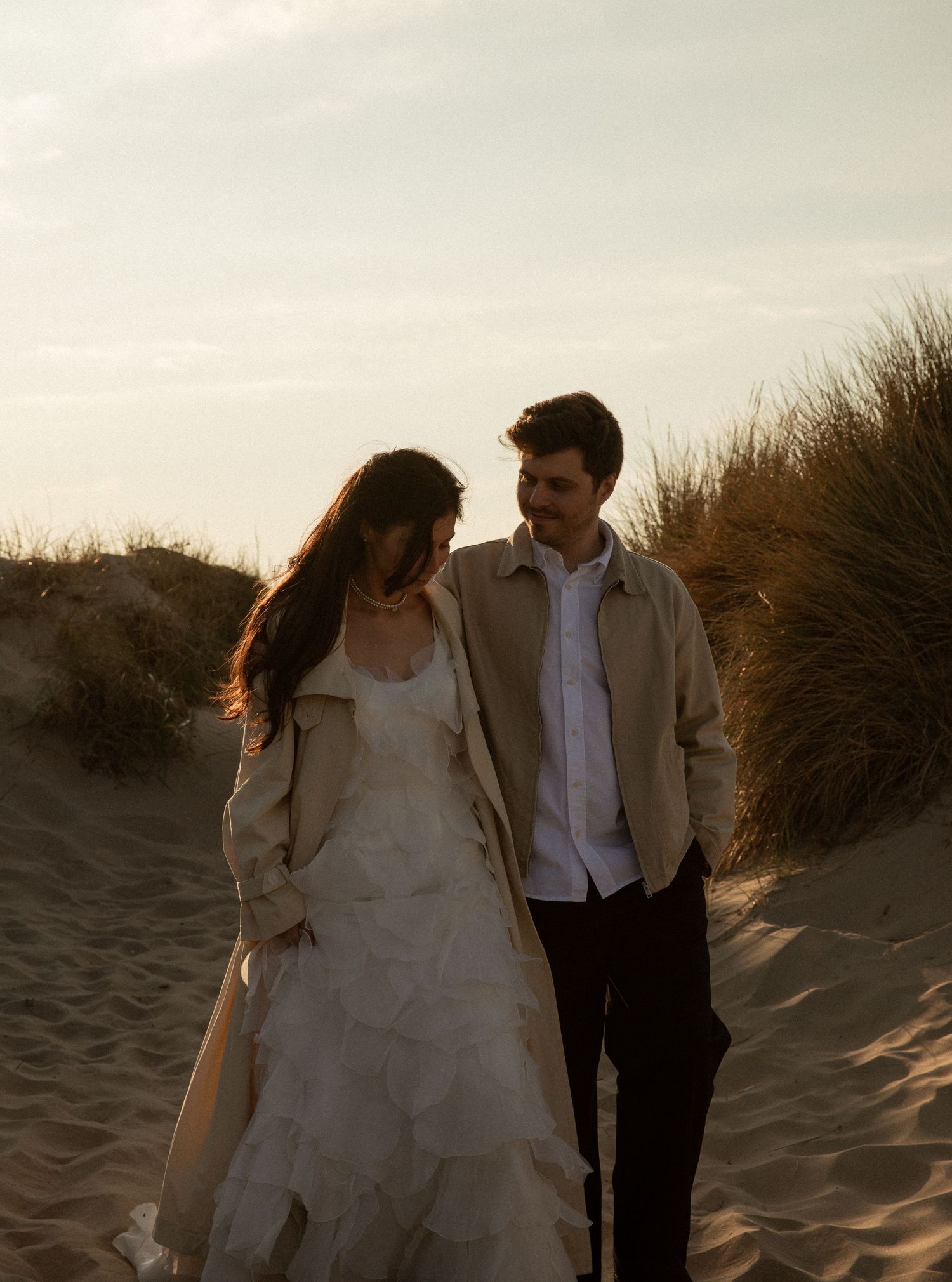 Couple walking on sandy beach dunes at sunset, woman in white ruffled dress with beige coat, man in tan jacket.
