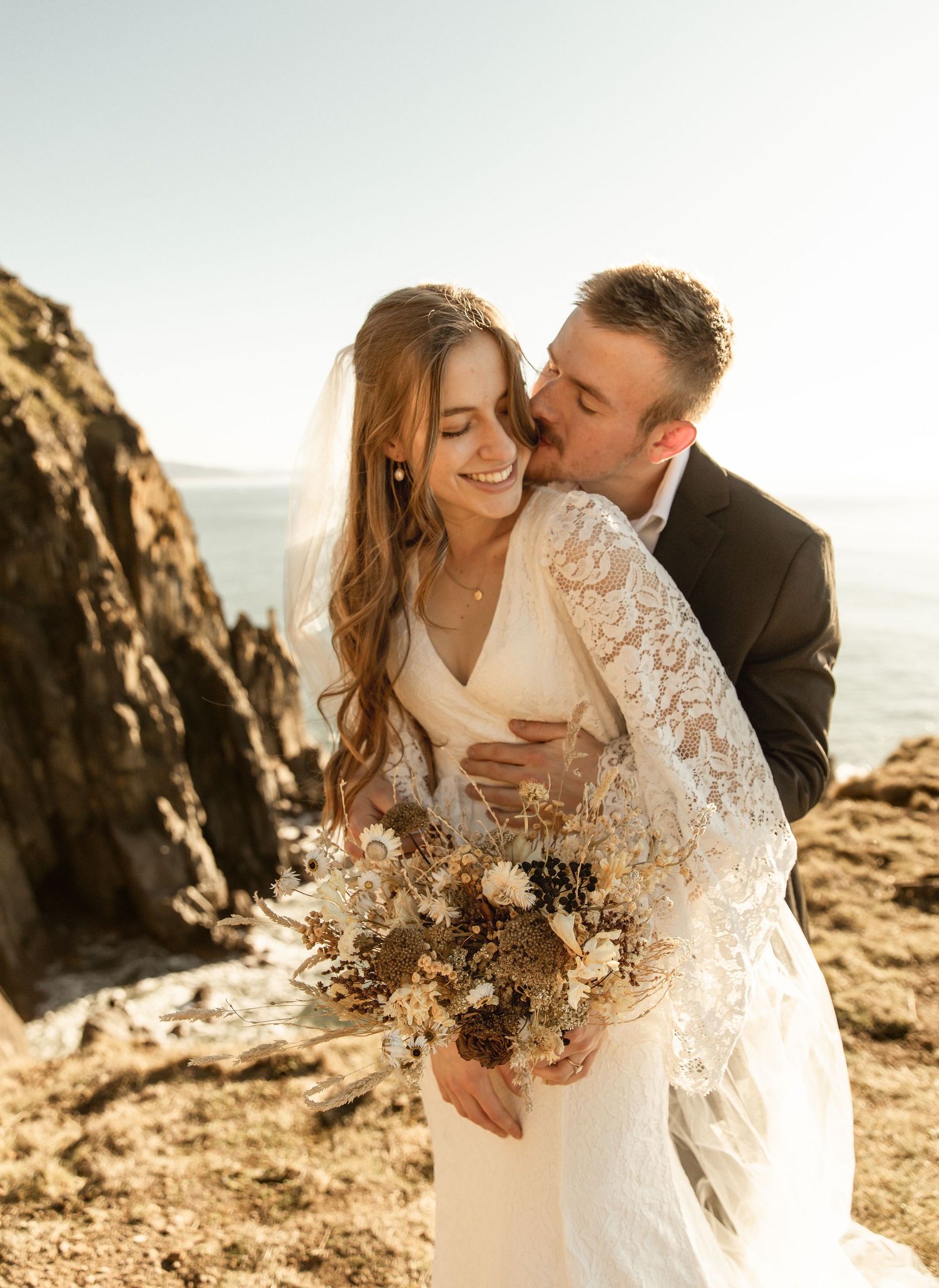 A romantic wedding couple embraces on coastal cliffs at sunset with a dried flower bouquet.