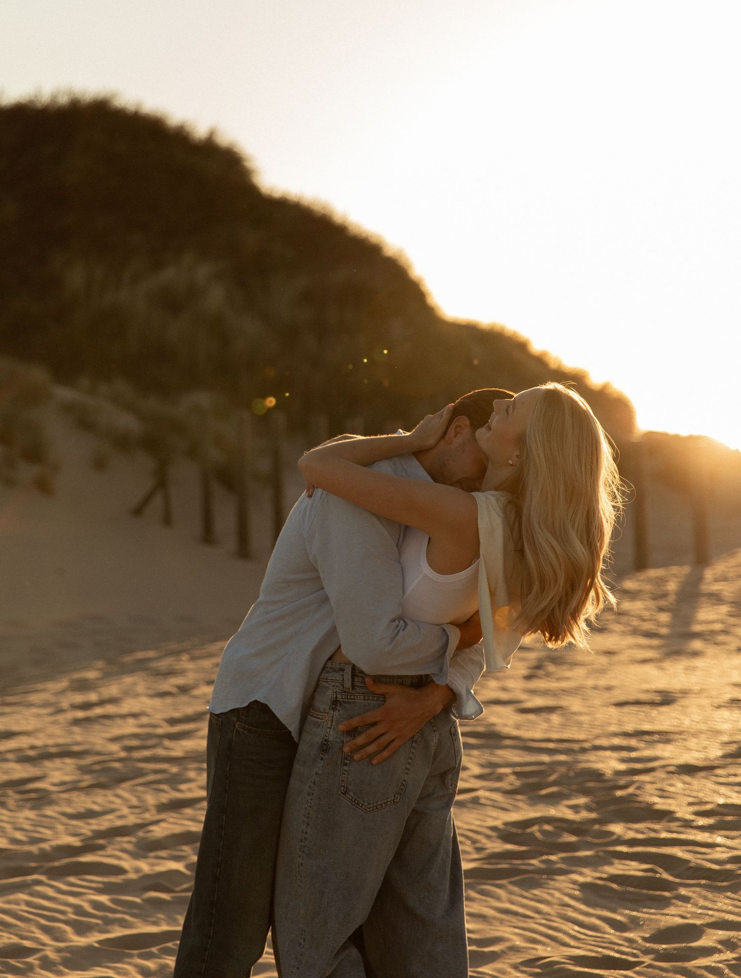 Couple sharing a romantic kiss on sandy beach dunes at golden sunset, woman in white top and jeans embracing partner.
