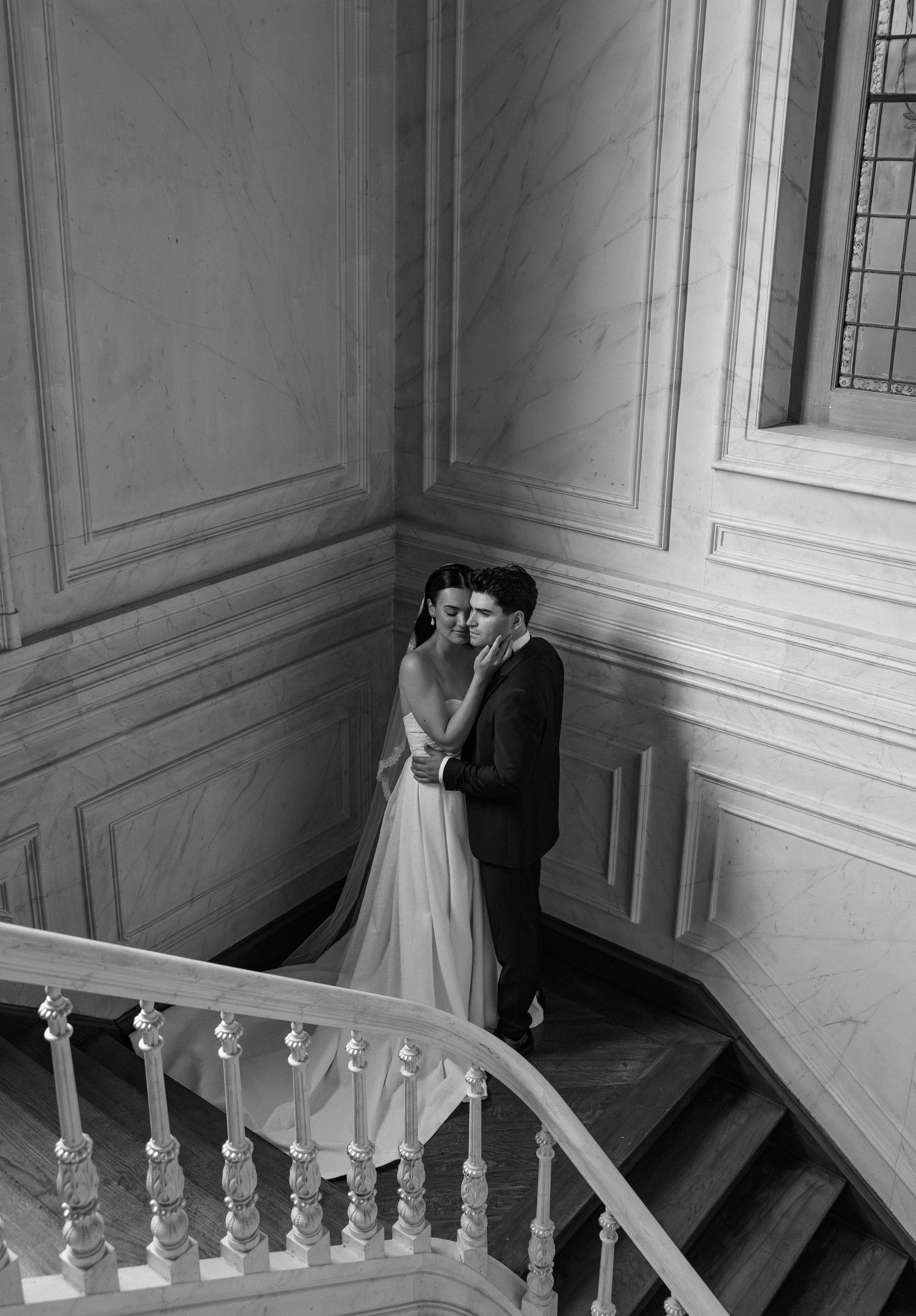 Black and white wedding photo of bride and groom embracing on elegant marble staircase in grand venue.
