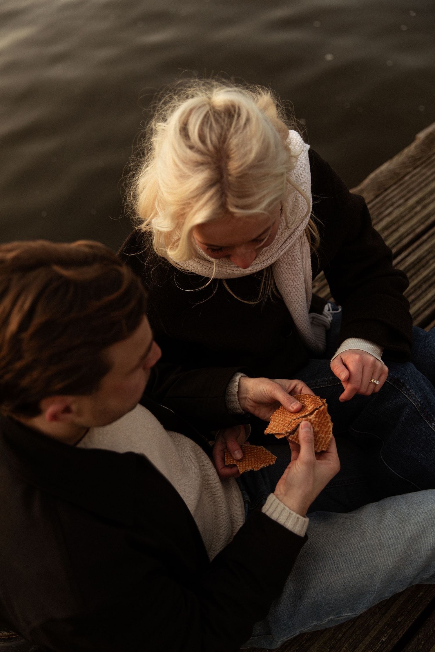 Two young people sitting on a wooden dock by the water, examining an autumn leaf together.