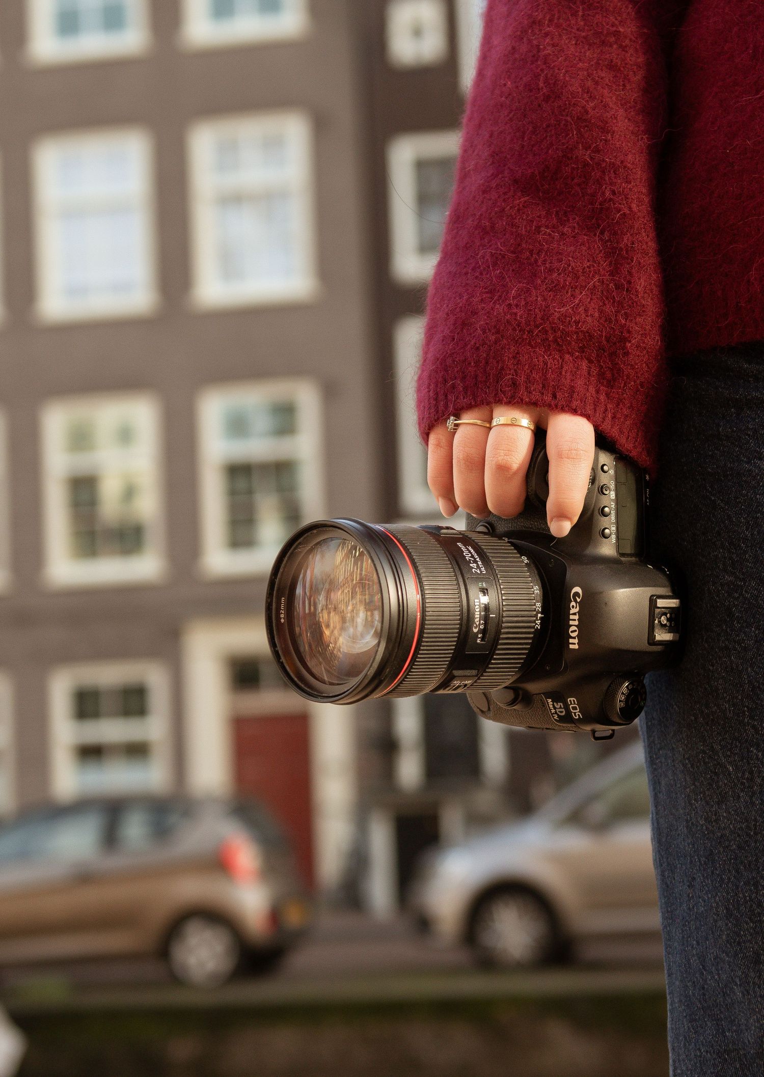 Person in red sweater holding a Canon DSLR camera with a zoom lens, standing in front of a blurred urban building.