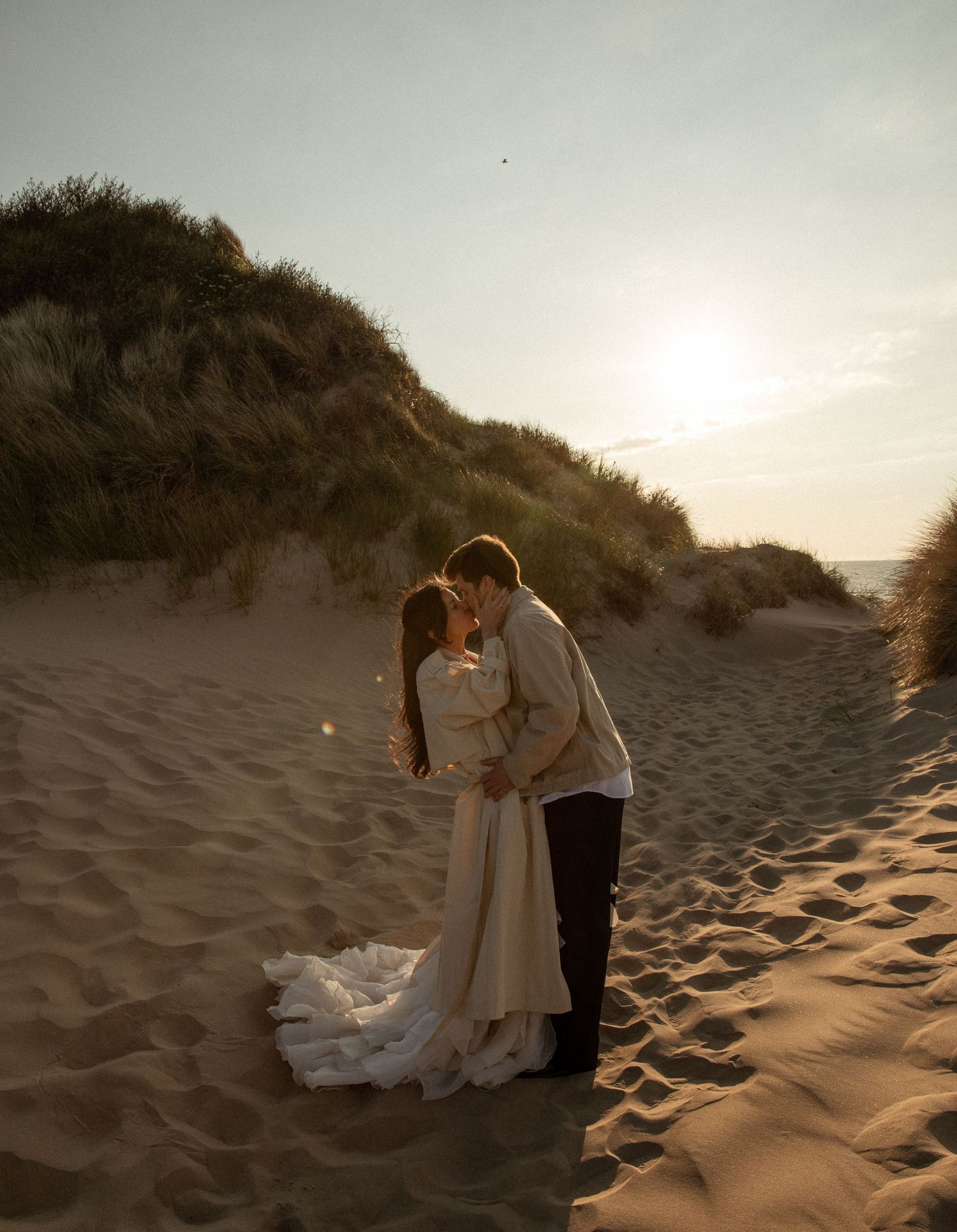 A couple shares a romantic kiss on sandy dunes at golden hour, bride in flowing white gown, groom in dark pants.