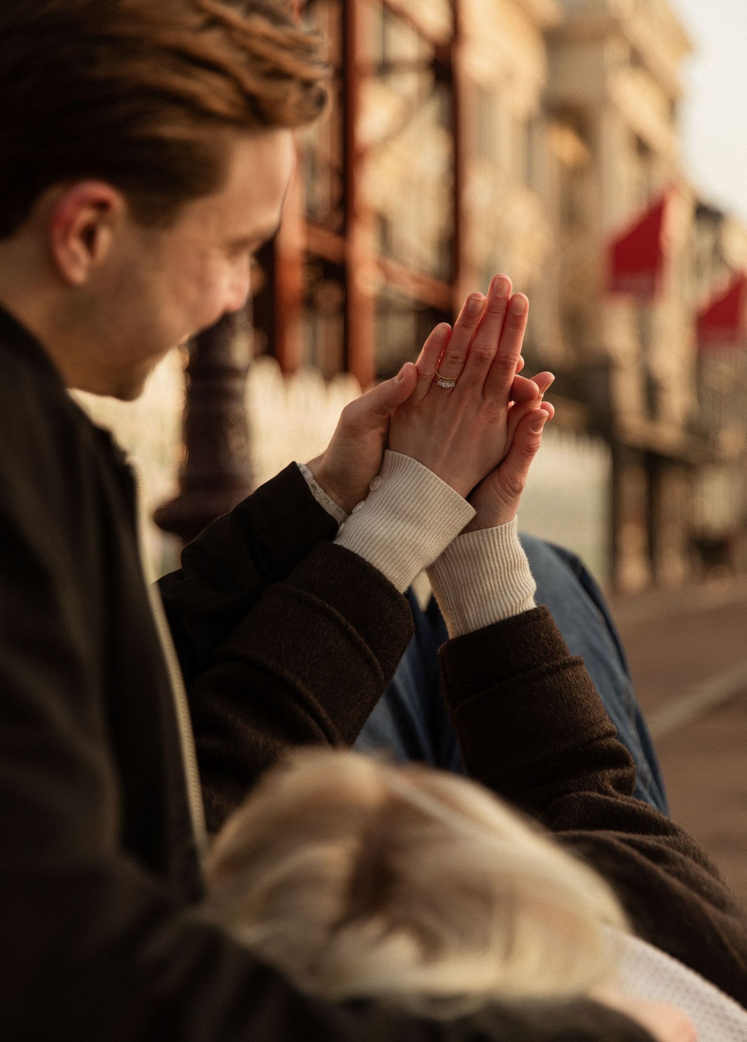 Two people press their hands together in prayer position on a city street with blurred architecture in the background.