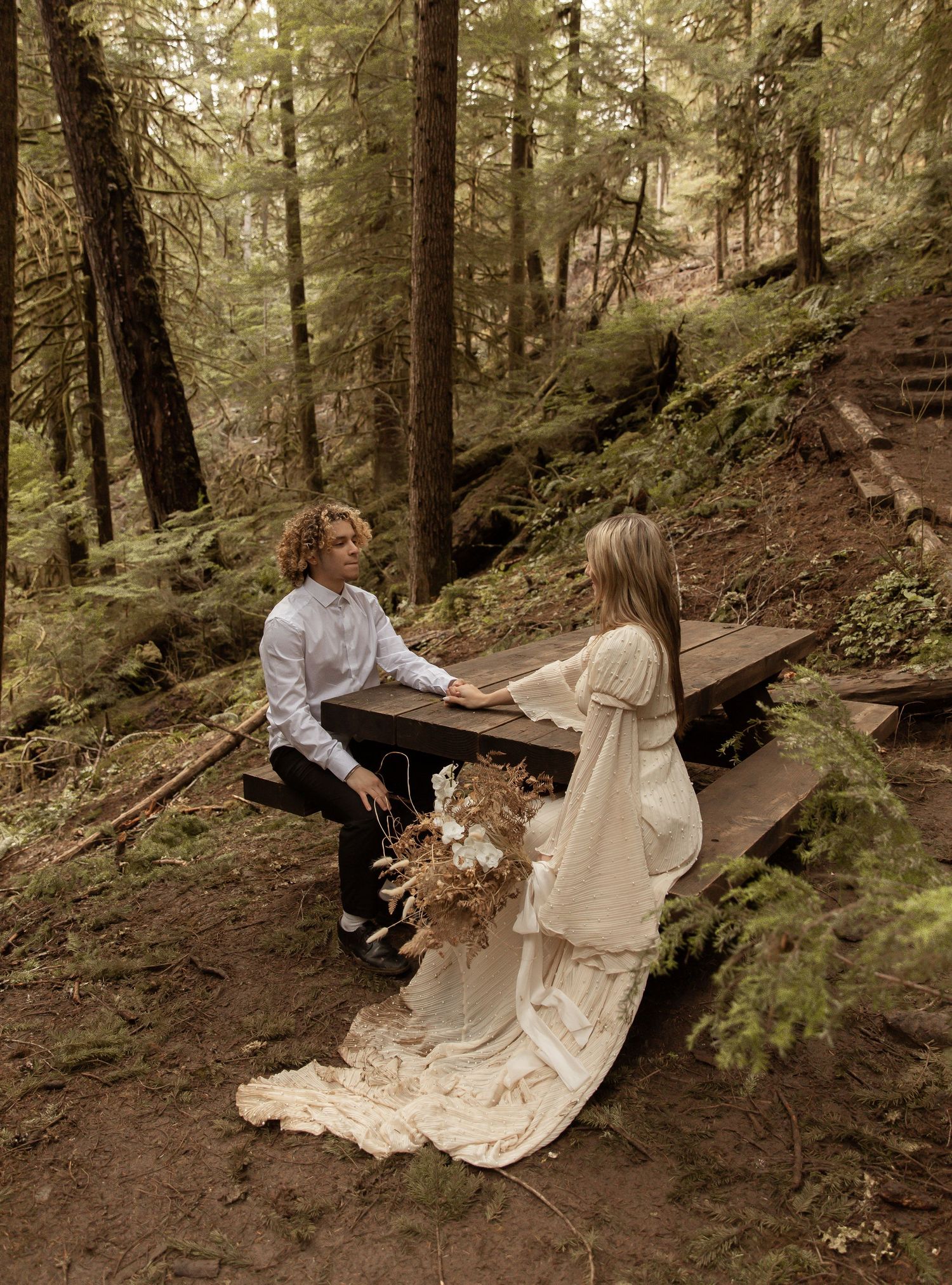 A couple sits on a wooden bench along a forest trail wearing formal attire.