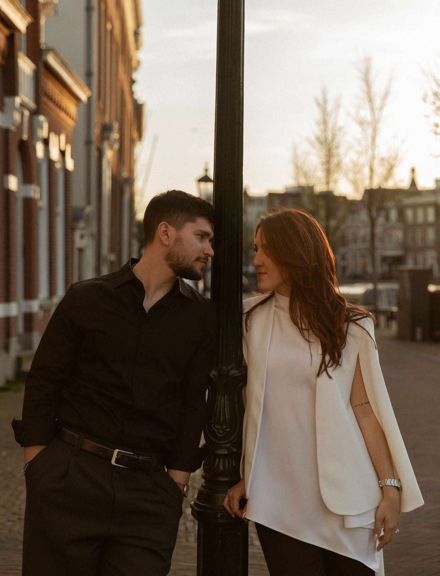 Couple gazing at each other by a lamppost on a golden-hour Amsterdam street, man in black shirt, woman in white cape top.