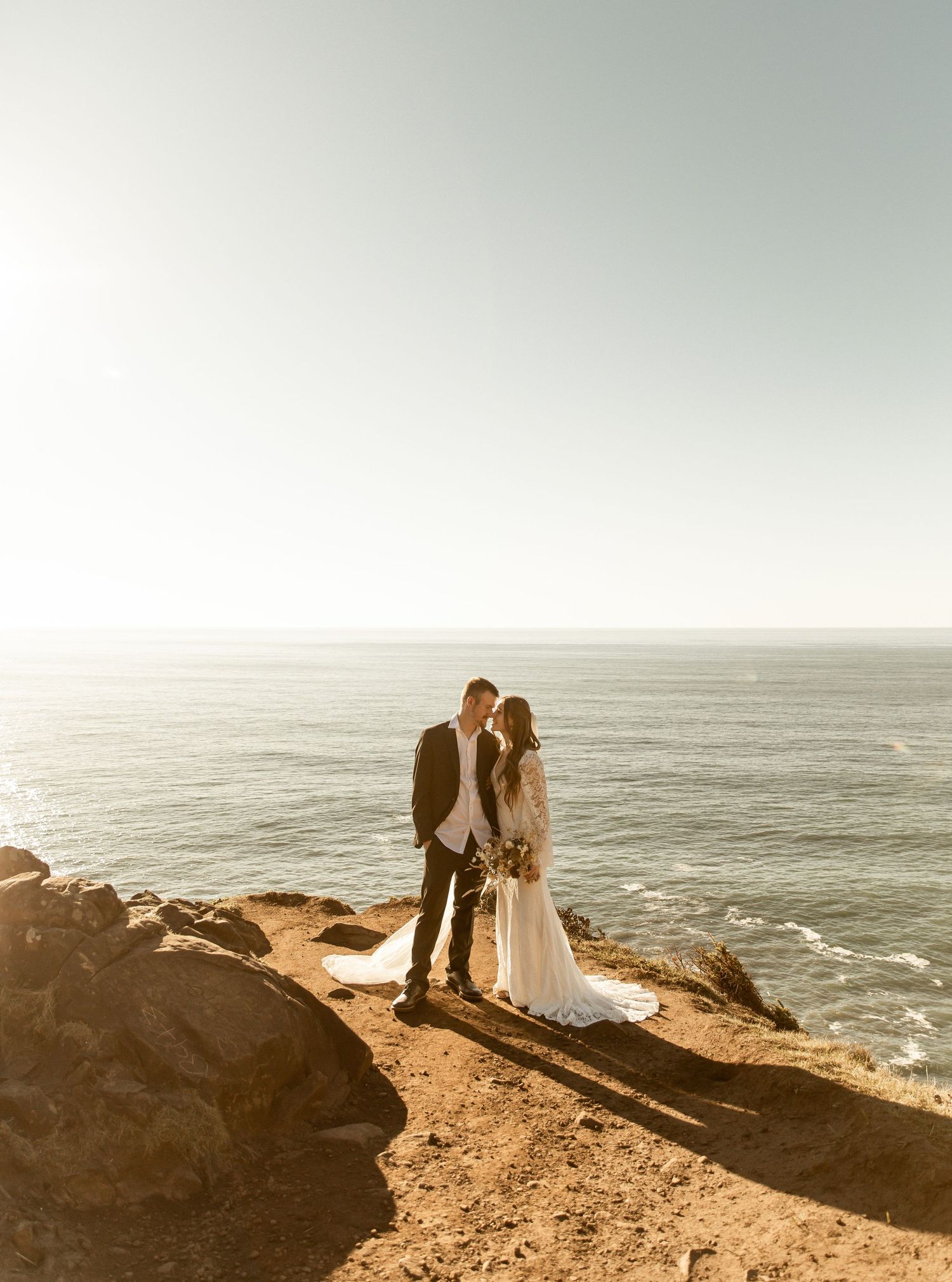 A coastal wedding photoshoot on dramatic cliffs overlooking the ocean at sunset.