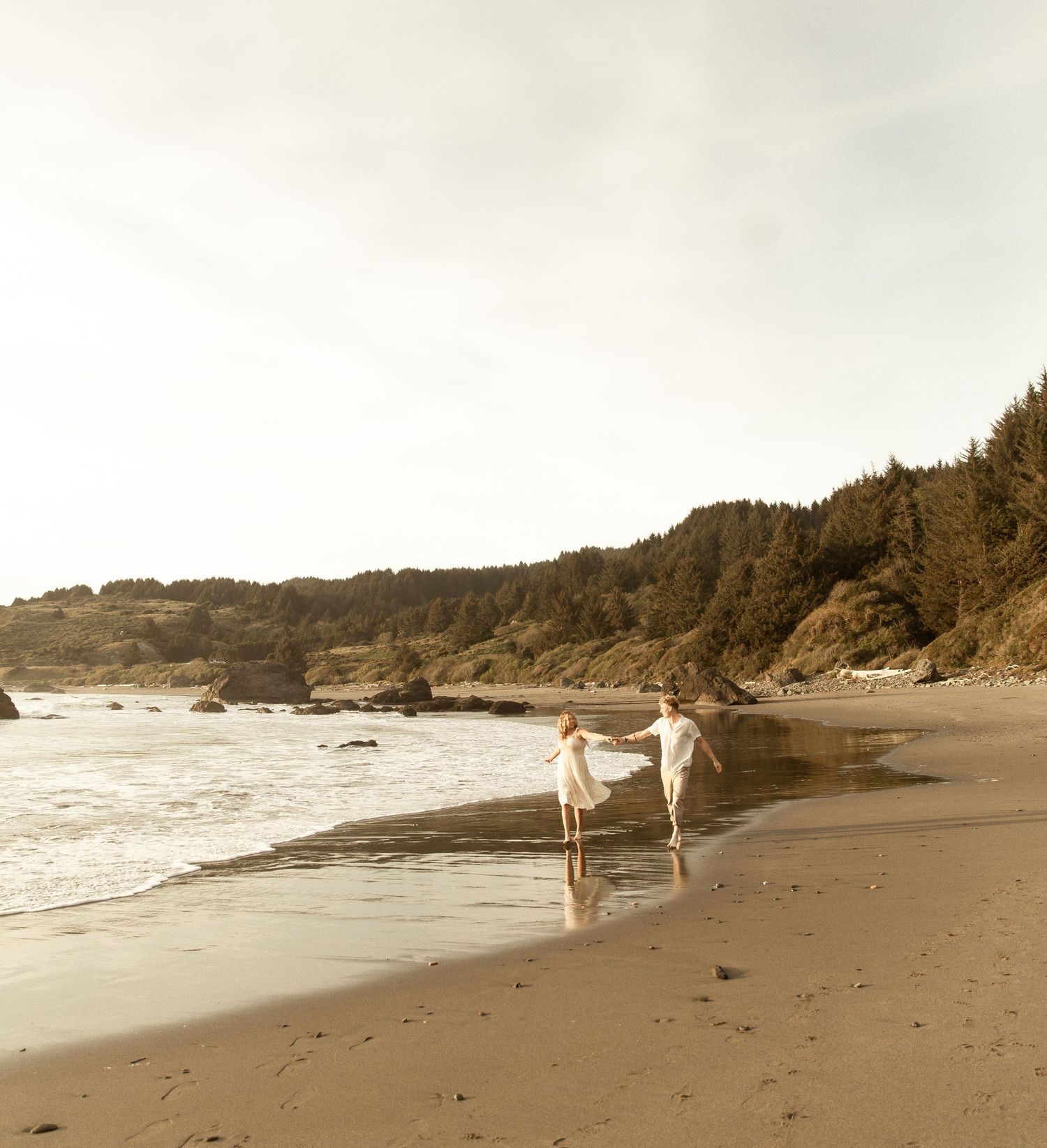 A panoramic view of a sandy beach with waves crashing on the shore and coastal cliffs in the background.