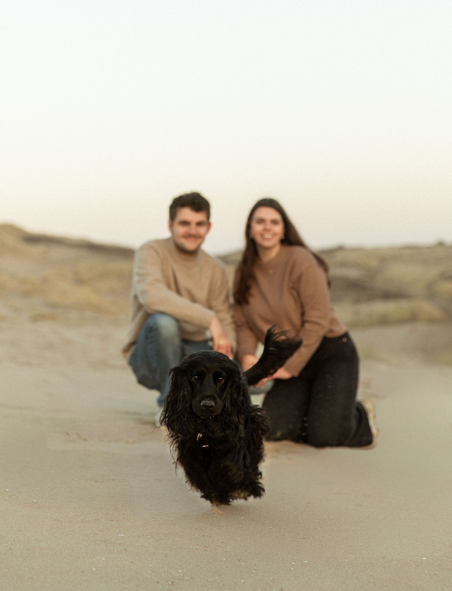 A couple relaxing on sand dunes with their black dog.