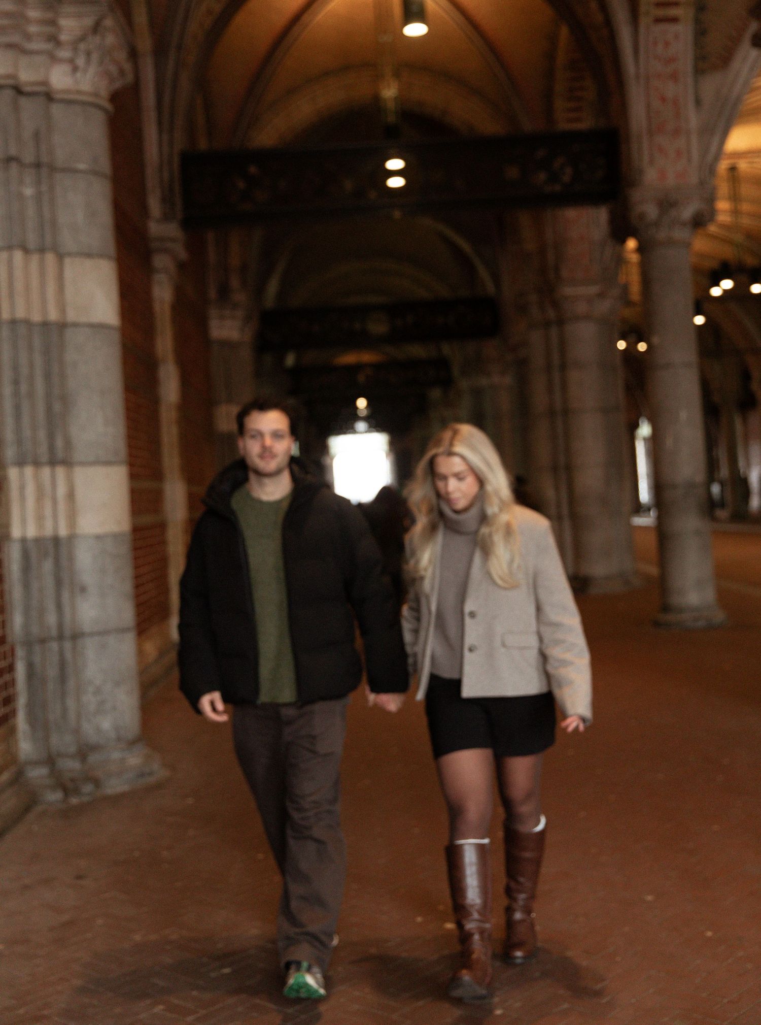 A couple walks together under an ornate archway with stone columns and warm lighting in a historic building.