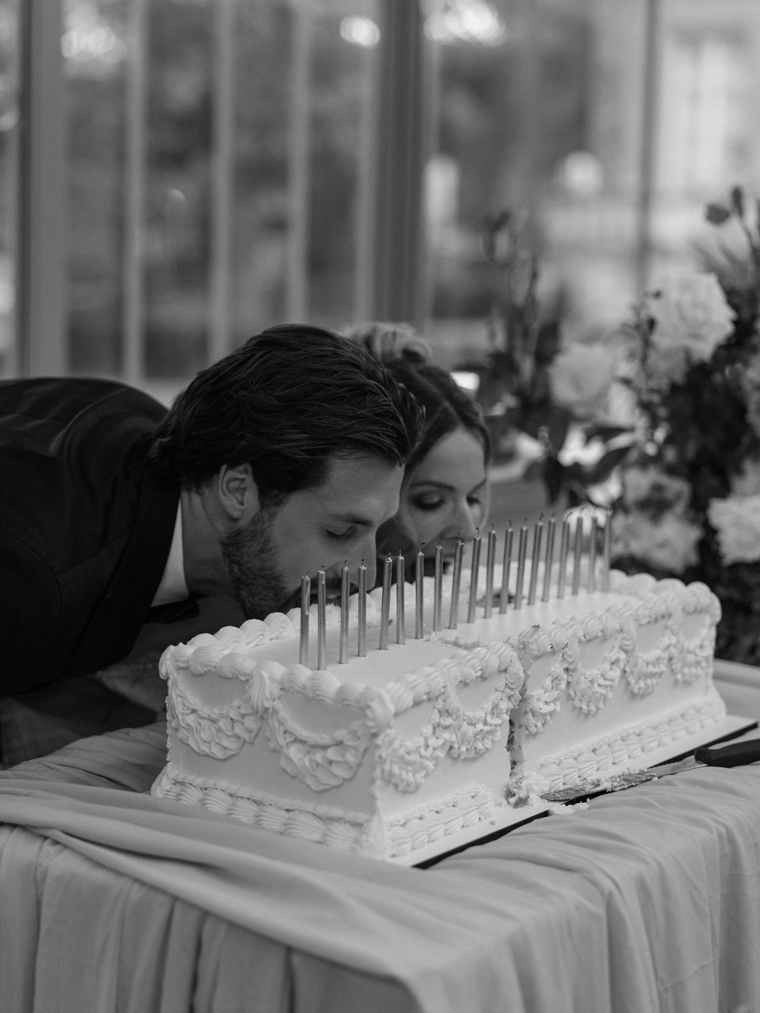 Couple blowing out candles on a white wedding cake in black and white photo at elegant reception venue.