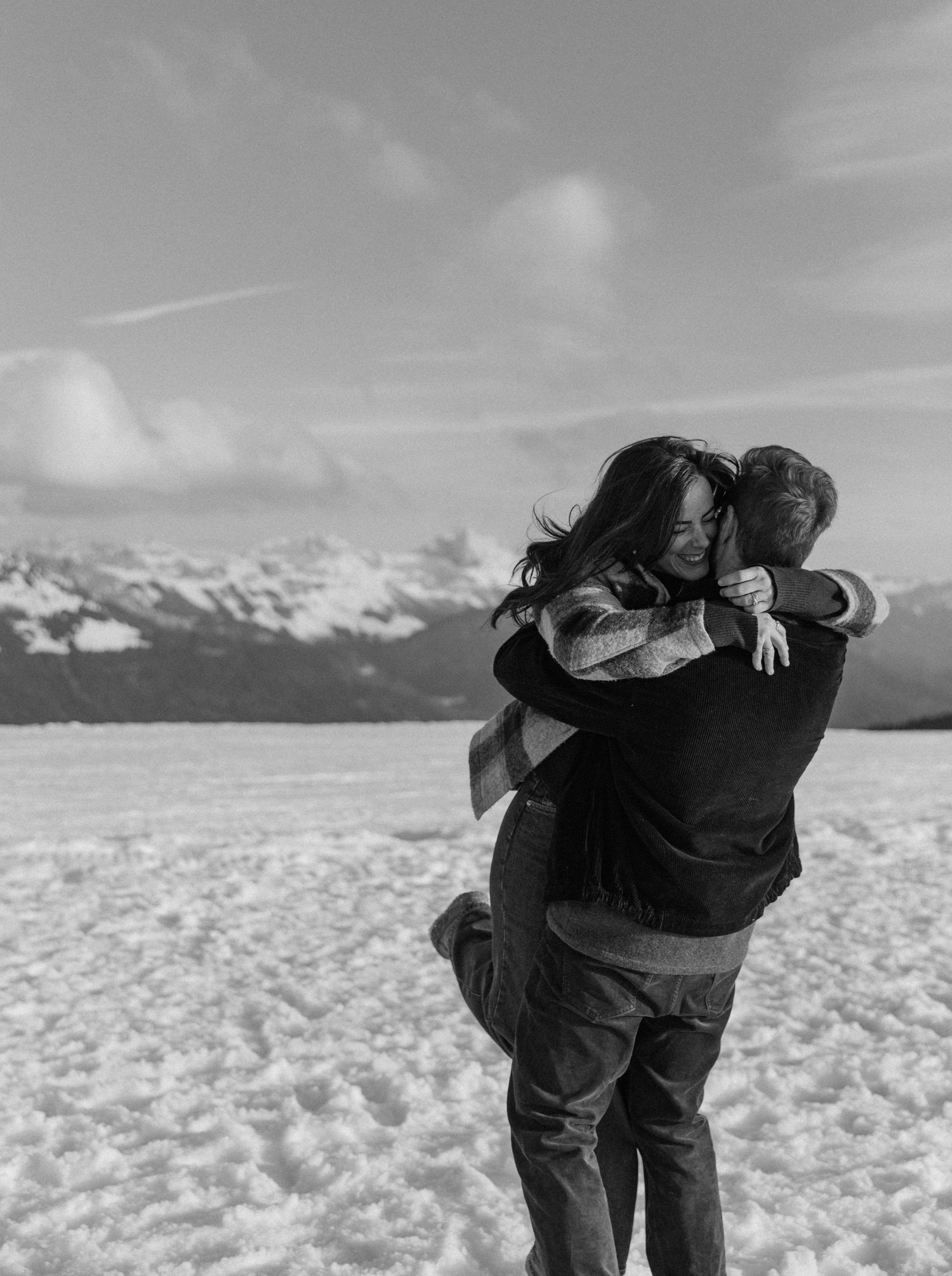 A couple embraces and spins together on a snowy mountain landscape under cloudy skies in a romantic winter moment.