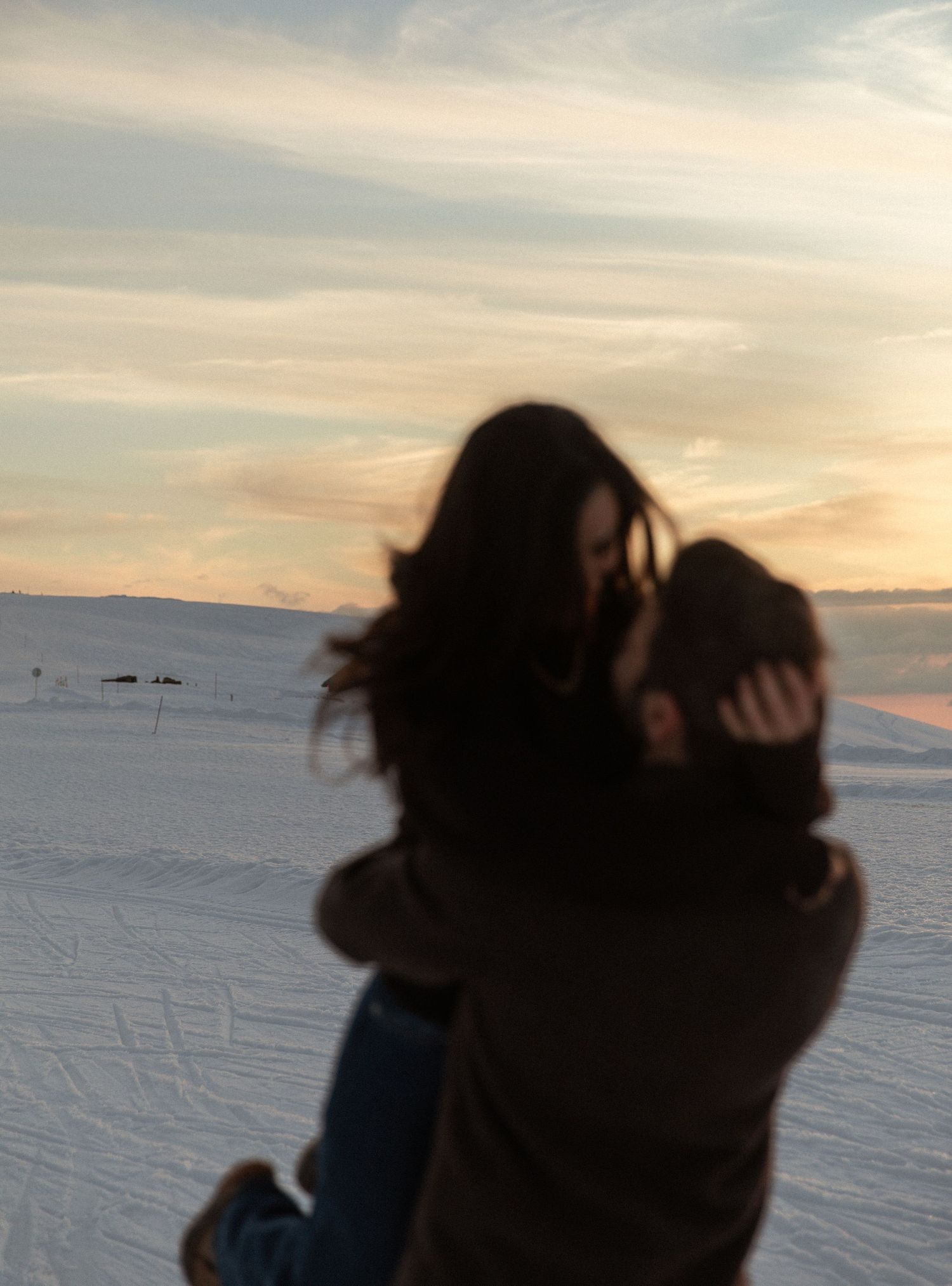 Silhouettes of people embracing against a snowy winter landscape at sunset with mountains in the distance.