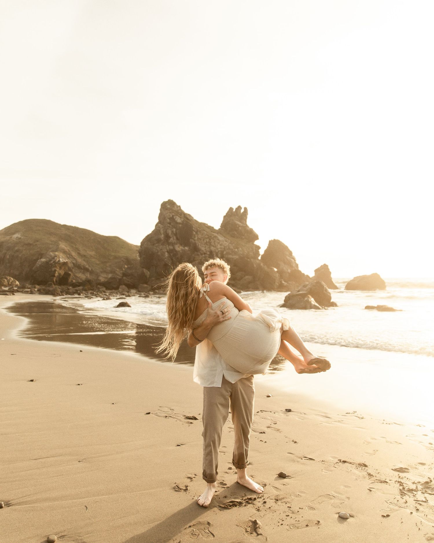 A couple strolls along a misty beach with rock formations in the background during golden hour.