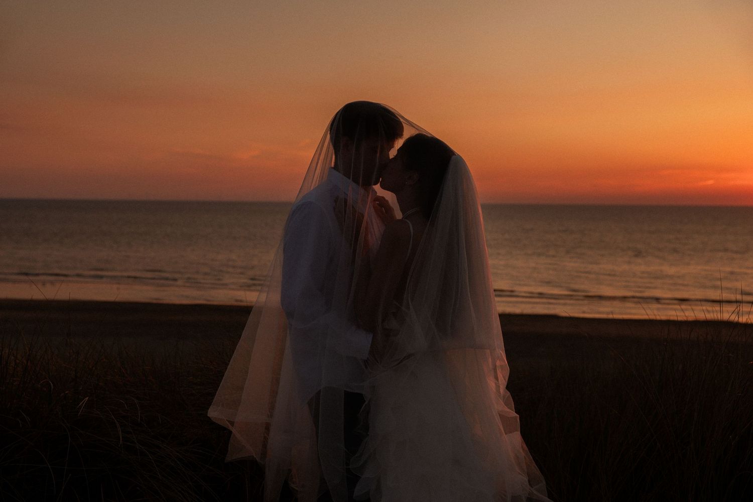 A bride and groom share a romantic kiss silhouetted against a stunning orange sunset over the ocean.