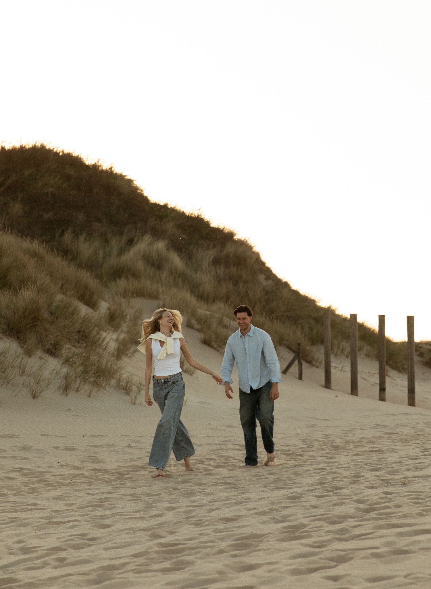 A couple walks hand-in-hand along a sandy beach dune path at sunset, with grass-covered hills and wooden posts behind them.