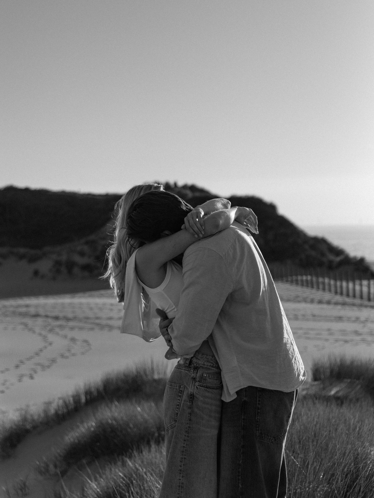 Black and white photo of a couple embracing and kissing on a sandy beach with dunes and hills in the background.