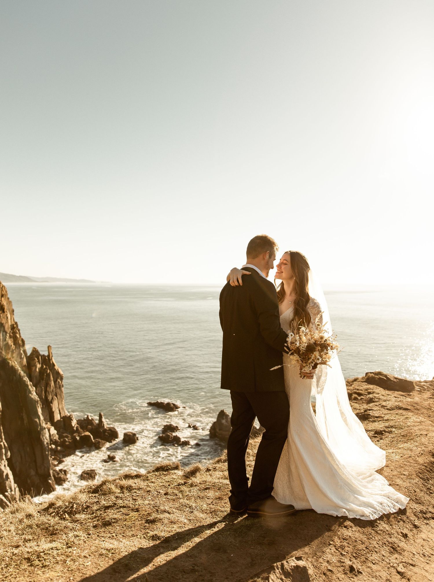 Wedding couple overlooking dramatic coastal cliffs at sunset.