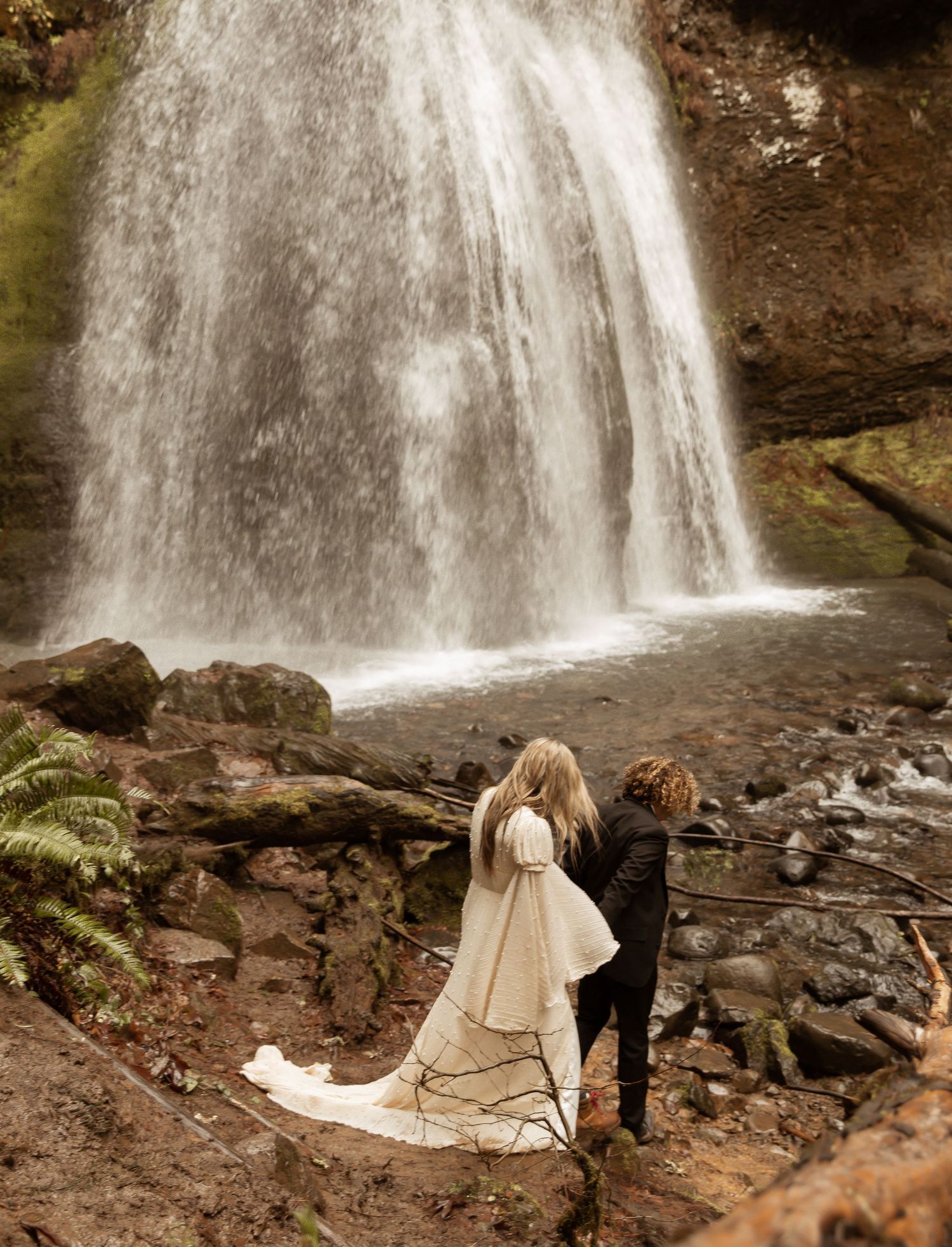 Two people in formal attire stand near a powerful waterfall surrounded by moss-covered rocks.