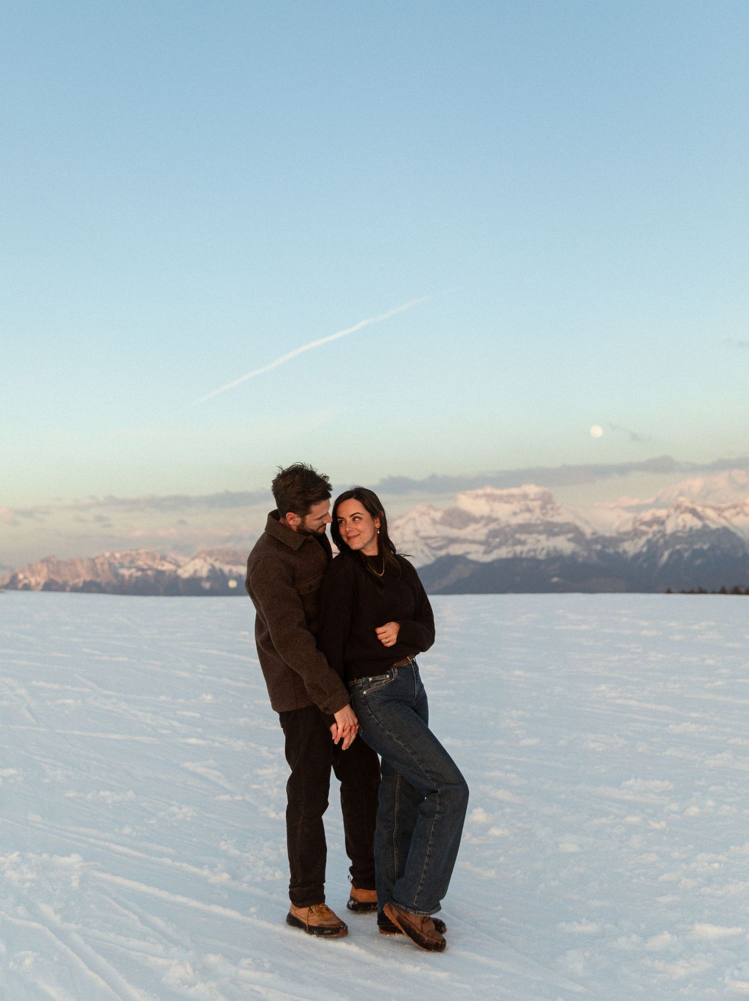 Two people stand together on a vast snowy field with snow-capped mountains visible in the distance during sunset.