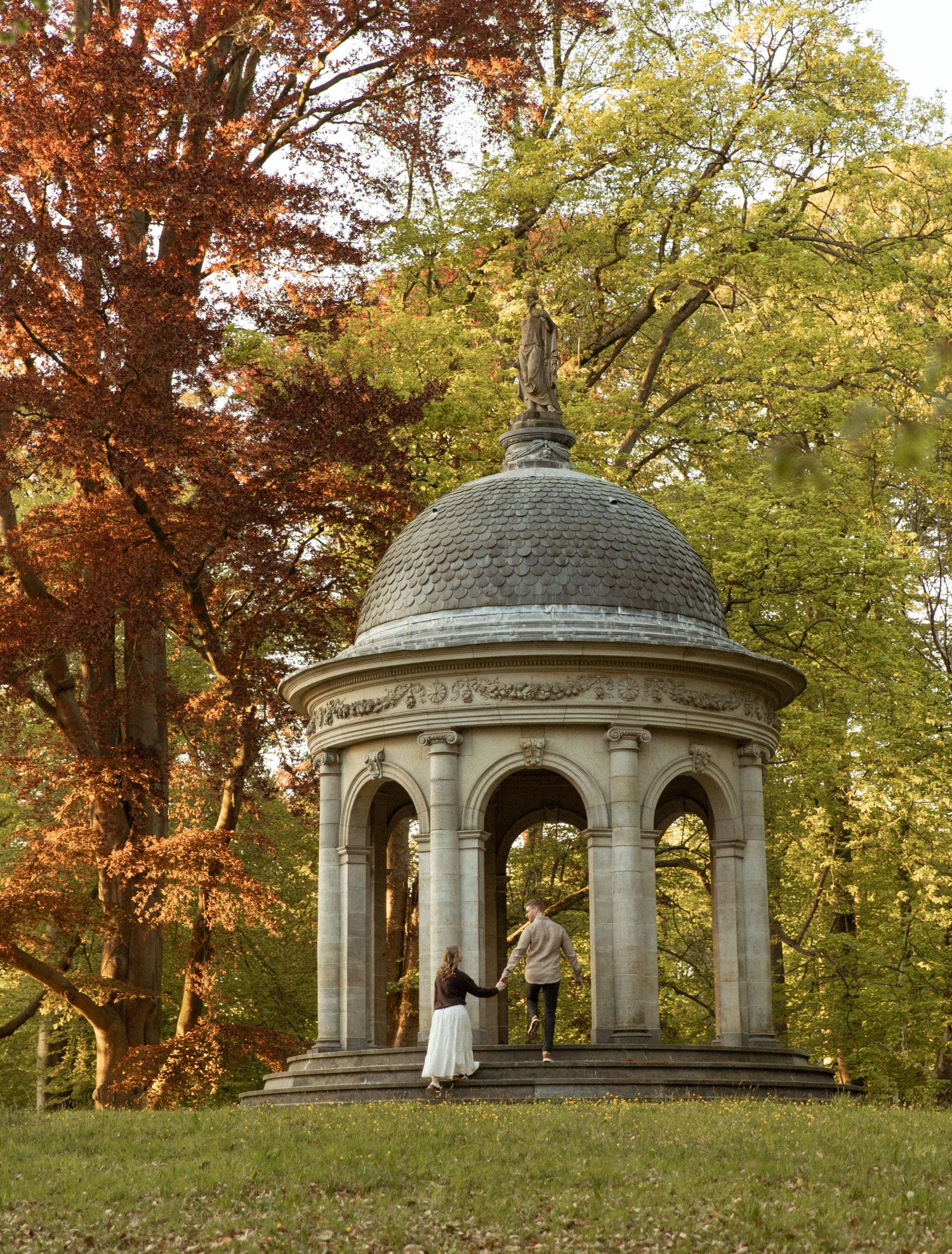 Couple exploring a classical stone gazebo with domed roof amid vibrant autumn foliage in a scenic park.