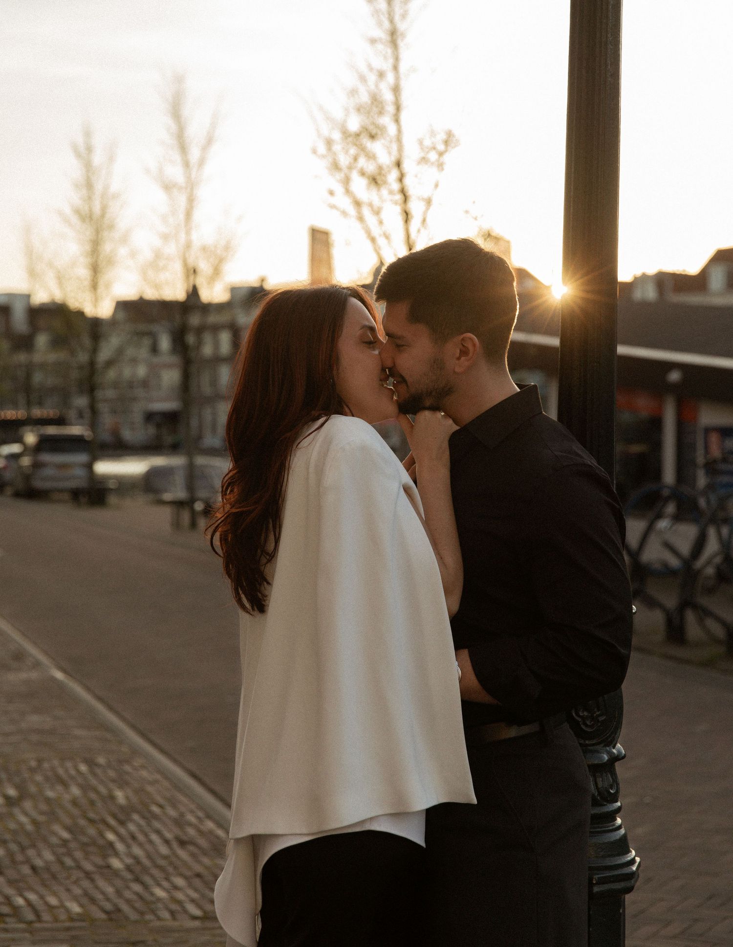 Couple sharing a romantic kiss at sunset on a cobblestone street with warm golden backlight.