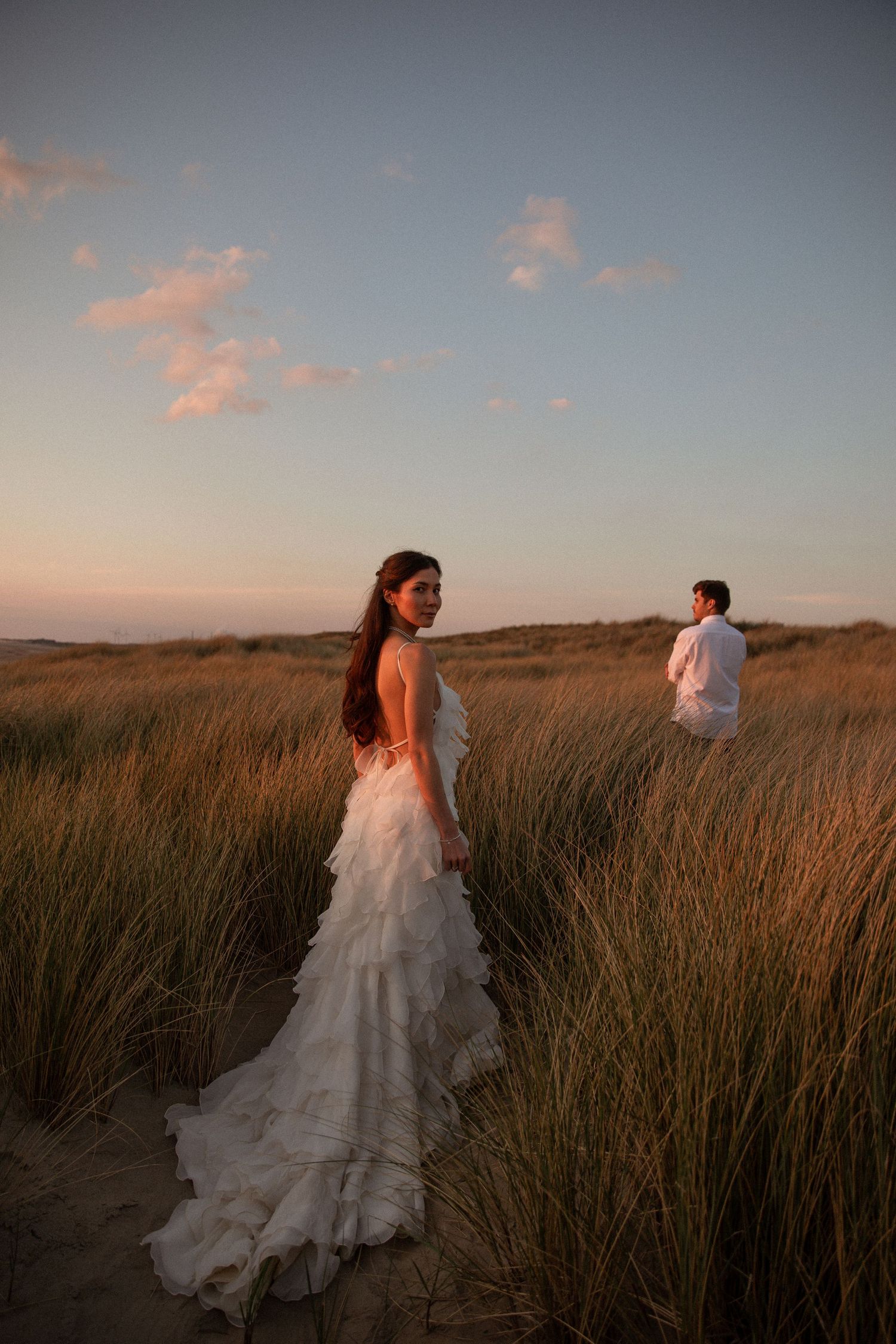 Bride in ruffled white gown stands in golden grass dunes at sunset, groom in white visible in background.