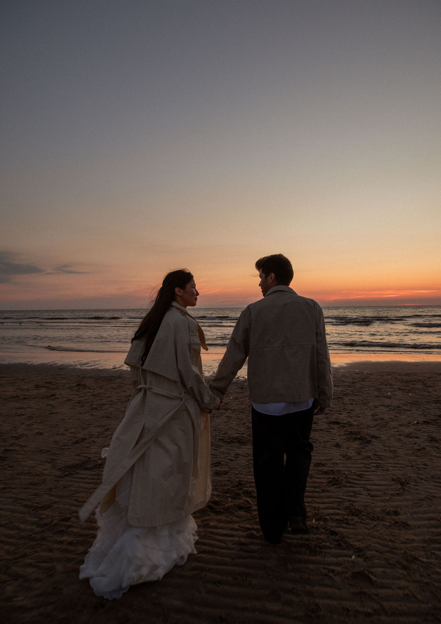 Couple holding hands on a sandy beach at sunset, facing the ocean in elegant formal attire.